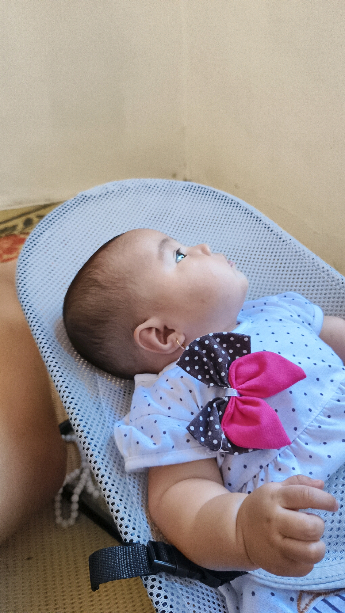 An infant wearing a white polka dot dress with a pink and gray bow is reclined in a gray baby bouncer. The baby is looking upwards with their head tilted. A small gold earring is visible on the baby's left ear. The background consists of a beige wall and a patterned rug. The scene appears to be indoors in a residential setting in Donorejo, Indonesia.
