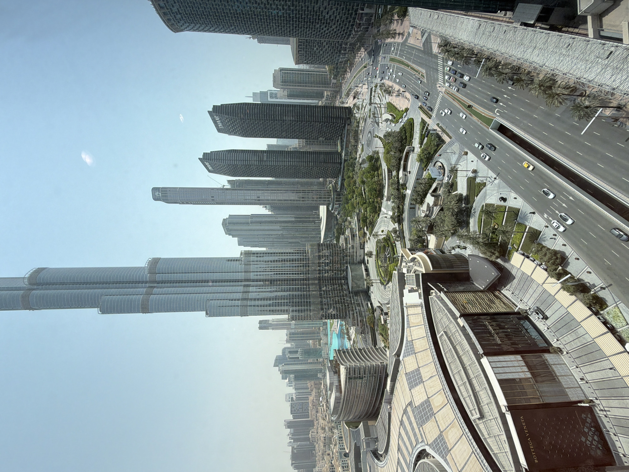 This is an aerial shot of downtown Dubai, United Arab Emirates, showcasing a modern urban landscape dominated by skyscrapers and an expansive mall. The Burj Khalifa, the world's tallest building, stands prominently on the left side of the frame, its sleek glass facade reflecting the clear blue sky. To the right of the Burj Khalifa, several other modern skyscrapers rise, some bearing the "EMAAR" logo.
In the foreground, the large, multi-level Dubai Mall is visible, characterized by its curved architecture and a vast, light-colored roof structure dotted with solar panels. The mall's facade features prominent luxury brand names like "BOTTEGA VENETA" and "DOLCE & GABBANA."
Surrounding the mall and the skyscrapers is a well-maintained urban environment with tree-lined streets, green spaces, and a large artificial lake or fountain area, likely part of the Dubai Fountain. Multiple lanes of traffic are visible on elevated roads and at ground level, with numerous cars moving along them. Palm trees are scattered throughout the landscape, adding a tropical touch to the cityscape. The overall impression is one of a bustling, contemporary metropolis under a bright, sunny sky.