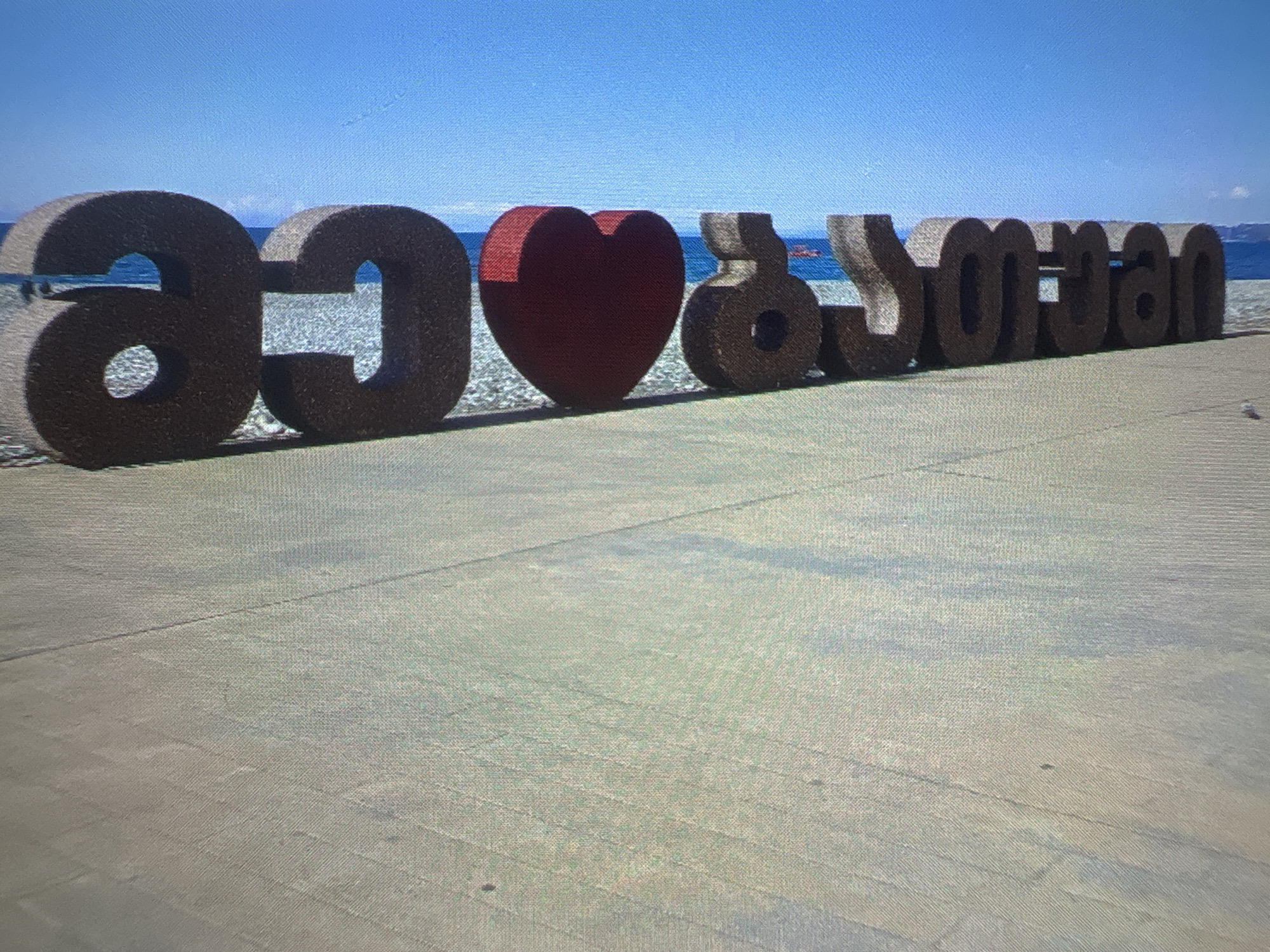 This is a close-up, vertically oriented photograph of a large, dark brown, three-dimensional sculpture spelling out "Batumi" with a red heart symbol in place of the letter 'u'. The sculpture is positioned on a textured, light-colored surface, likely a promenade or boardwalk, next to a body of water that appears to be the sea, with a clear blue sky above. The water is visible behind the sculpture, with a hint of a person in the distance swimming or wading. The lighting suggests it is daytime, with some shadows cast by the sculpture. The texture of the sculpture is rough, possibly made of concrete or a similar material. The overall scene conveys a coastal, possibly tourist-oriented, location.