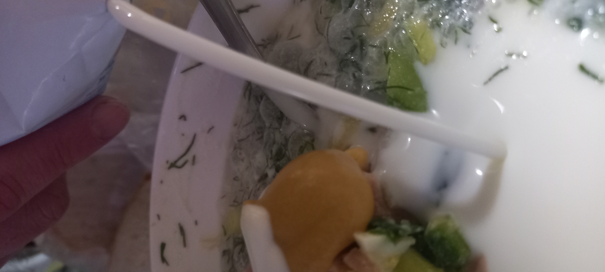 A person is pouring a white liquid from a carton into a white bowl. The bowl contains a light-colored soup with green herbs and pieces of yellow and green vegetables. A spoon is visible in the bowl. The image is taken from a close-up perspective, with the hand and carton in the upper part of the frame. The location is not discernible from the image.