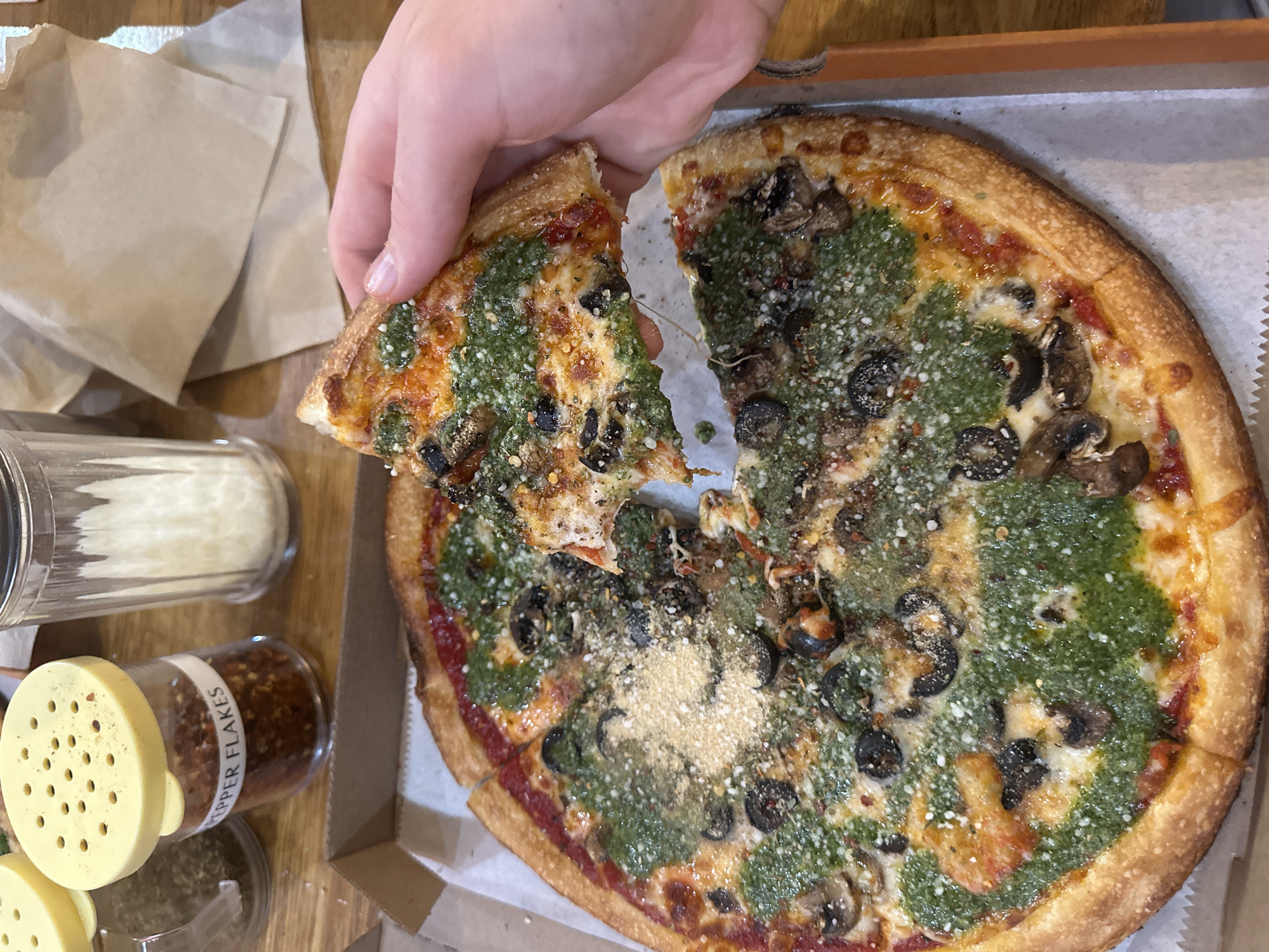 An overhead shot captures a person's hand lifting a slice of pizza from a whole pizza, which rests in a cardboard box on a light brown wooden table. The pizza features a golden, slightly thick crust and is topped with a blend of red tomato sauce and a generous drizzle of vibrant green pesto. Black olive slices and mushrooms are scattered across the surface, along with visible sprinkles of grated white cheese and red pepper flakes. As the slice is pulled away, strands of melted cheese stretch between the pieces, indicating the pizza is warm and freshly served.
To the left of the pizza, two seasoning shakers are visible. A tall, clear glass shaker contains white, grated contents, likely parmesan cheese. Below it, a smaller shaker with a yellow cap and a clear body holds red pepper flakes, with the label "PEPPER FLAKES" clearly readable. A brown paper napkin is also seen crumpled on the table in the upper left. The setting suggests a casual dining experience, possibly in College Park, USA.
