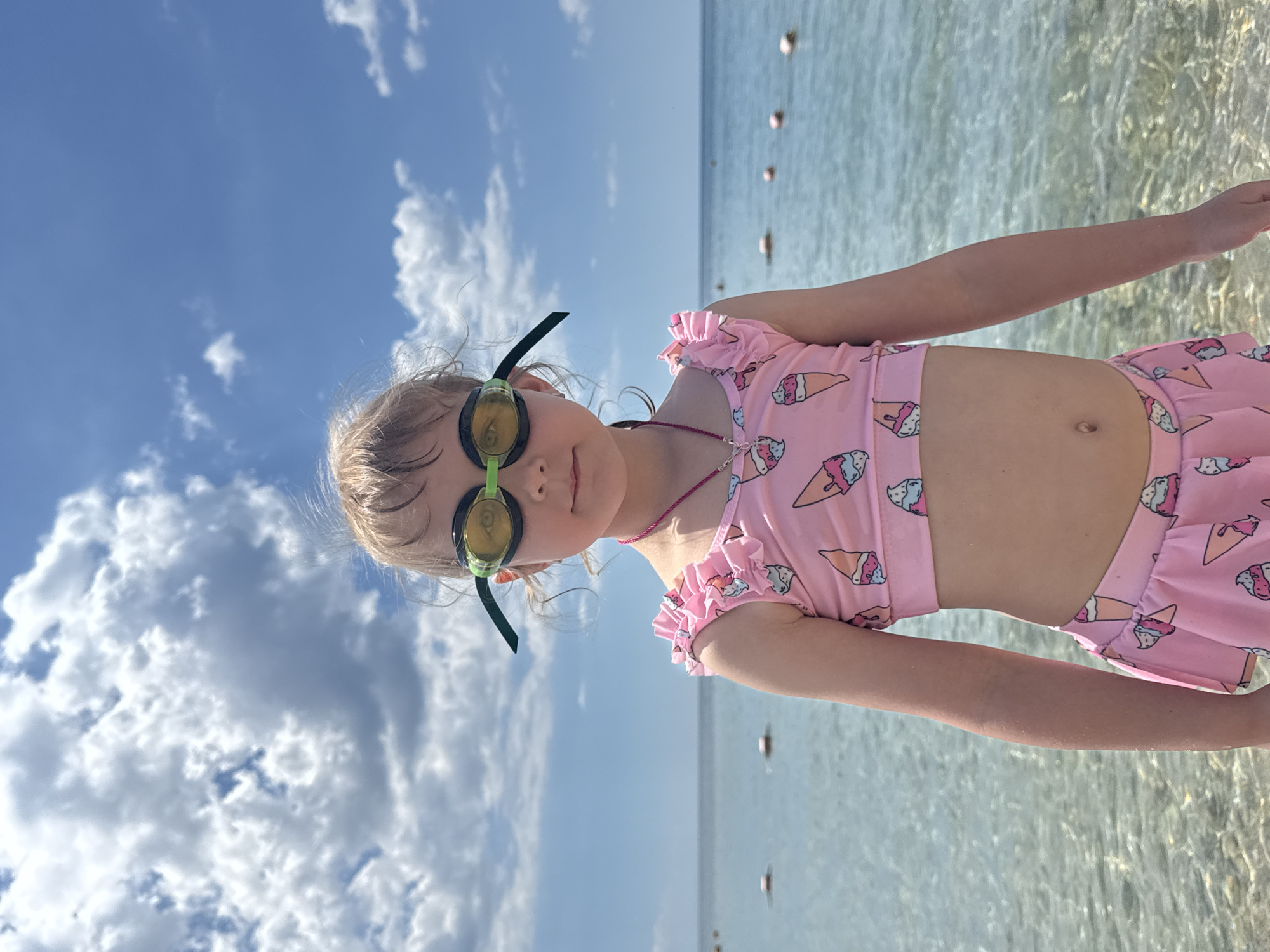 A young girl stands in shallow, clear water. She wears swimming goggles and a pink swimsuit with an ice cream cone pattern. The sky above is blue with scattered white clouds. In the background, a string of buoys marks the edge of a swimming area. The image was likely taken at a beach in Yevpatoriya, Ukraine.