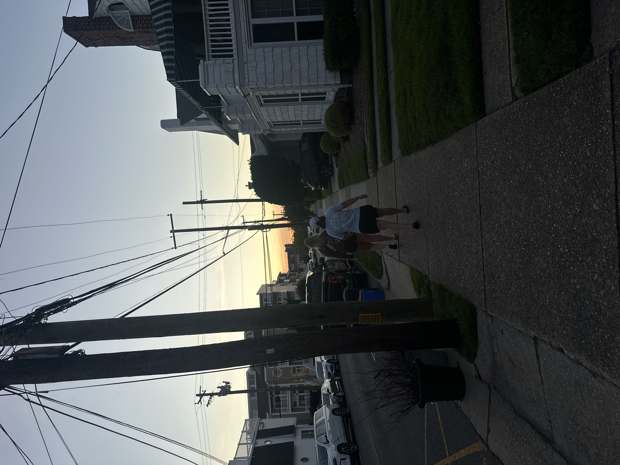 Two individuals are walking south on a sidewalk in Ocean City, United States of America, during twilight. The individual in the foreground is wearing a white cap, a light blue t-shirt with graphics, and dark shorts. The individual behind them is wearing a dark t-shirt and dark shorts. They are walking away from the camera. The sidewalk is made of concrete and is cracked in several places. To the left of the sidewalk is a street with parked cars and a black pickup truck. To the right of the sidewalk is a well-maintained lawn. In the background, there are houses and utility poles with numerous wires crisscrossing overhead. The sky is a gradient of blue and orange, indicating the sun has recently set. A white house with green awnings and a brick chimney is visible on the right side of the frame. The overall scene depicts a quiet residential street at dusk.