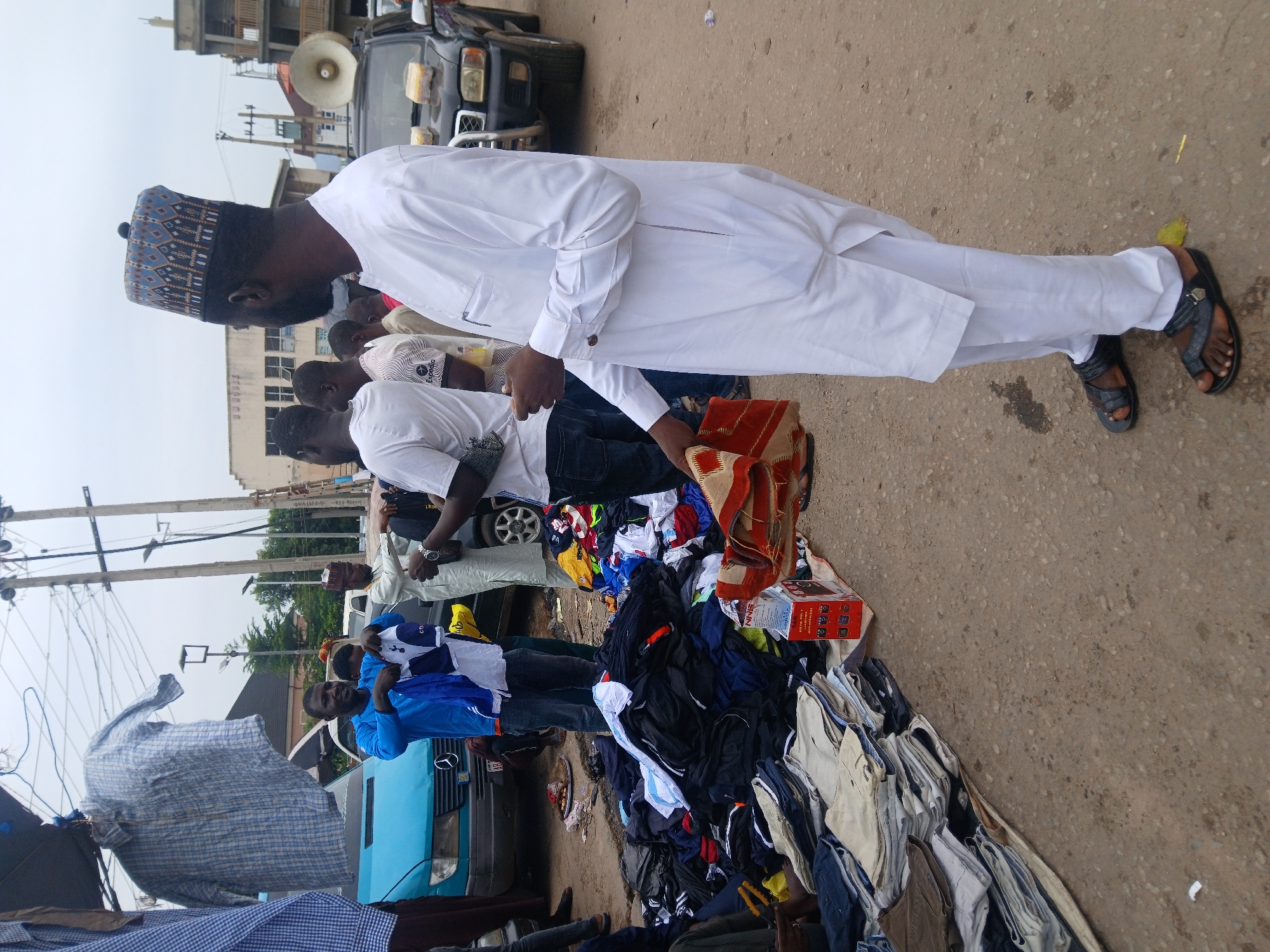 A street market scene in Abuja, Nigeria, shows a group of people gathered around a large pile of clothing spread on the ground. A man in a white traditional outfit and a patterned hat is interacting with the clothes. The scene features other individuals, a light blue van, a dark SUV equipped with a loudspeaker, and a variety of garments. Overhead electric lines are visible.