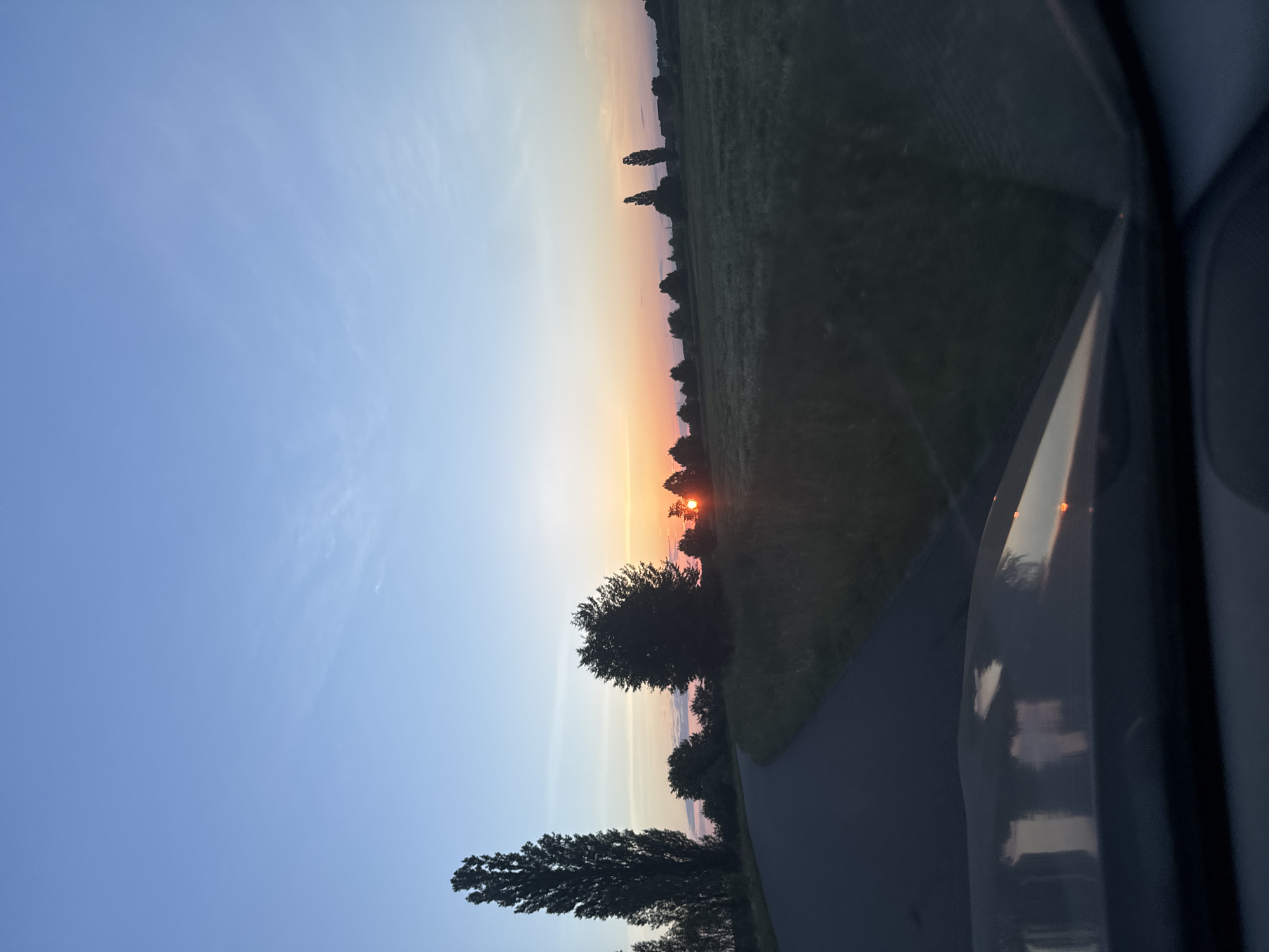 The image depicts a scenic sunset over a rural landscape in Dovhe, Ukraine. The sun is low on the horizon, casting a warm, orange glow across the sky, transitioning to soft hues of pink and yellow. The sky is a clear, pale blue with faint wisps of clouds. Silhouetted against the vibrant sky are several trees, their dark forms standing out starkly. A road or path curves through the landscape, bordered by a grassy field. In the foreground, part of a vehicle is visible, suggesting the photo was taken from inside a car. The reflections on the vehicle's surface hint at the surrounding environment, possibly other vehicles or the sky. The overall mood is peaceful and serene, capturing the beauty of a country sunset. The time of day is clearly evening, and the weather appears to be clear.