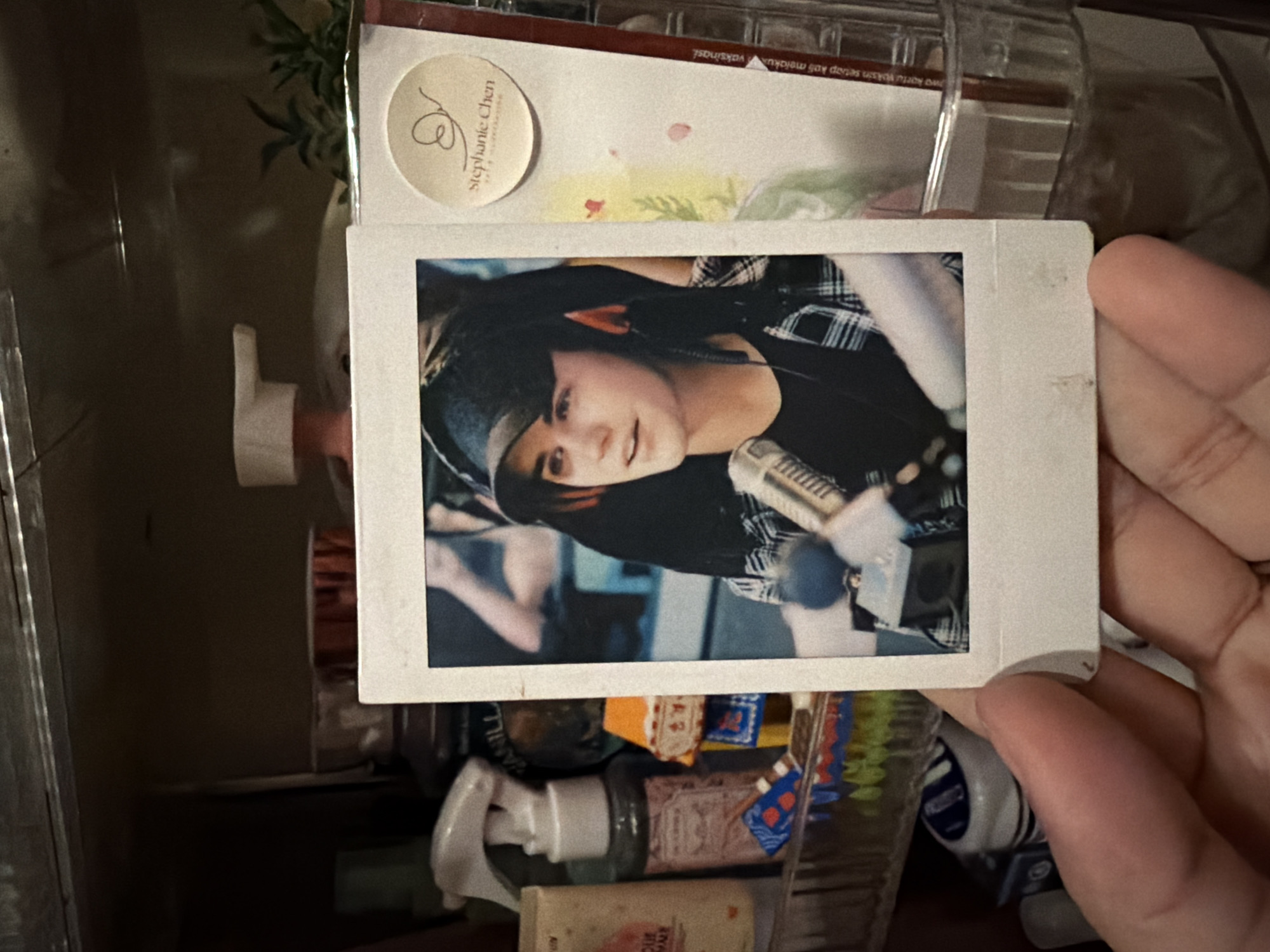 A Polaroid-style photograph depicting a person with dark hair and a black headband, wearing a black shirt and a plaid shirt, and standing in front of a microphone, is being held by a hand. The photo is taken indoors, above a clear plastic storage container filled with various items. The background suggests an interior space with additional storage containers and bottles visible. The image is located in East Jakarta, Indonesia.