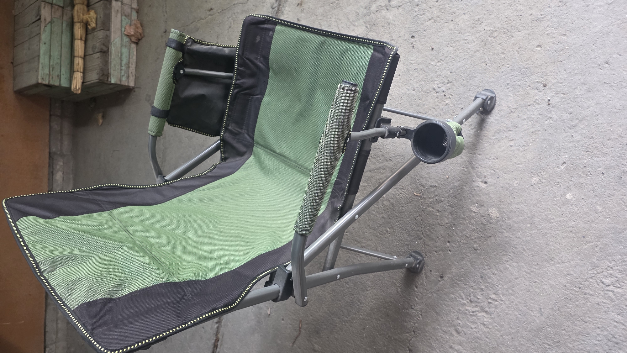 An empty green and black camping chair with a cup holder is set up on a concrete surface. The chair is made of fabric stretched over a metal frame. In the background, there are some wooden planks stacked on top of each other. The environment appears to be outdoors, possibly a workshop or an industrial area given the concrete floor and the stacked lumber. There are no people or animals present in the image. The scene does not offer any specific clues about its location within the city of Chuy, Kyrgyzstan, beyond the general description of an outdoor, utilitarian space.