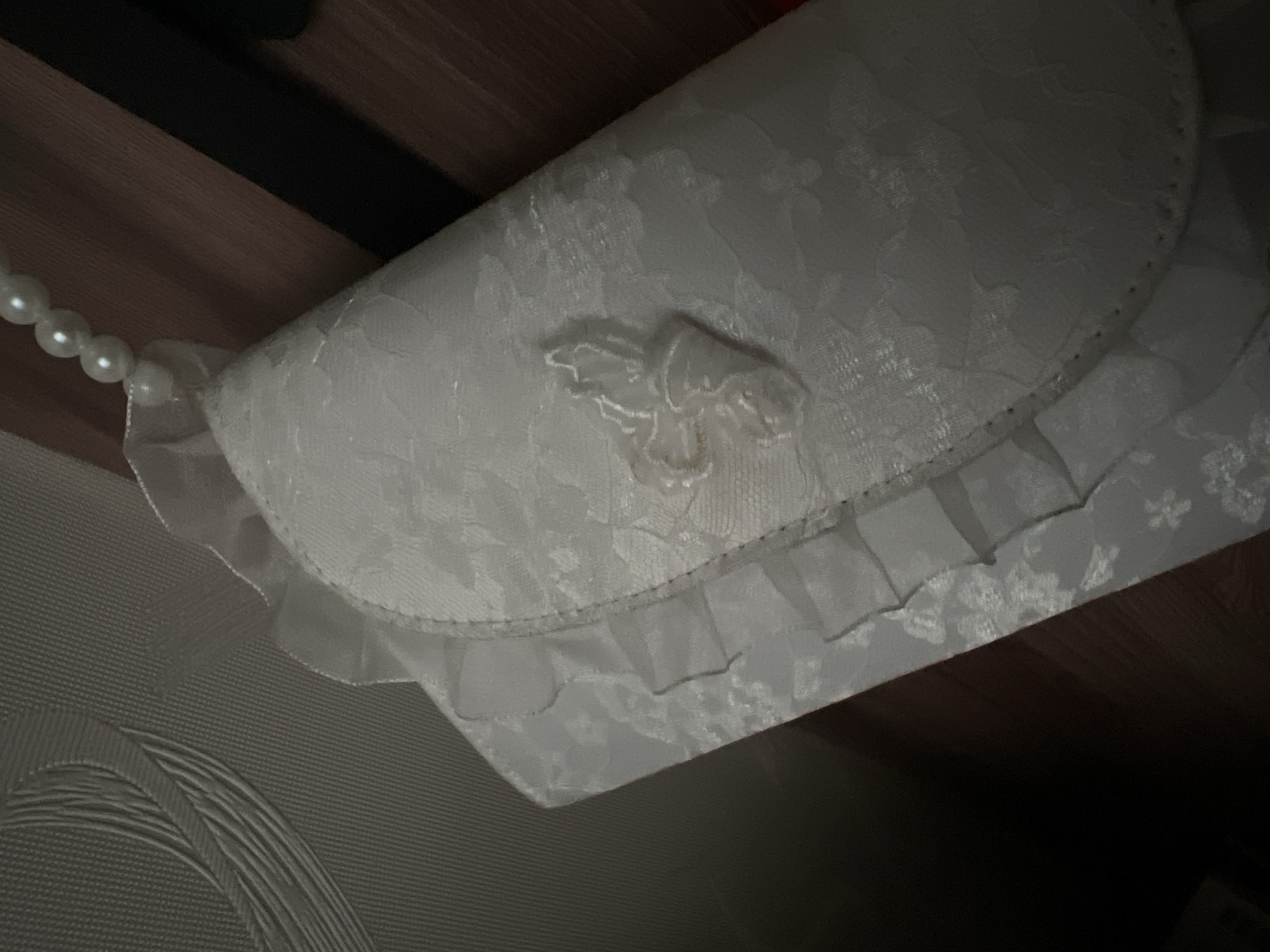 An ornate, white lace clutch purse with ruffled edges and a beaded strap is displayed against a wooden surface, likely a table or shelf. The purse features a textured floral pattern and a raised applique of a delicate flower. The background is dimly lit, with some texture visible on the wall above the purse. The overall impression is one of elegance and delicate craftsmanship. The image does not contain information about the city of Vetluzhsky, Russia.