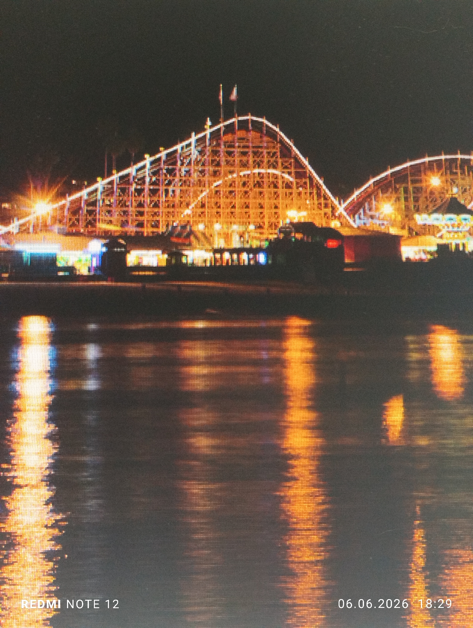 The image presents a nighttime scene of an amusement park in Burgas, Bulgaria, viewed across a dark body of water. The upper half of the frame prominently features multiple wooden roller coaster structures, extensively outlined and illuminated with warm orange and yellow lights. Two flags are visible at the apex of the leftmost roller coaster. Situated beneath the roller coaster tracks are various smaller park buildings and kiosks, emitting a combination of orange, blue, and white light. A structure resembling a carousel is discernible on the right side of the amusement park area. The lower half of the image captures the dark surface of the water, which reflects the amusement park's lights as elongated, shimmering streaks, predominantly in orange and yellow hues with some blue accents. The entire image displays a noticeable pixelated or grid-like visual artifact. Overlaid text at the bottom left reads "REDMI NOTE 12," and at the bottom right, "06.06.2026 18:29." No human subjects are discernible within the frame.