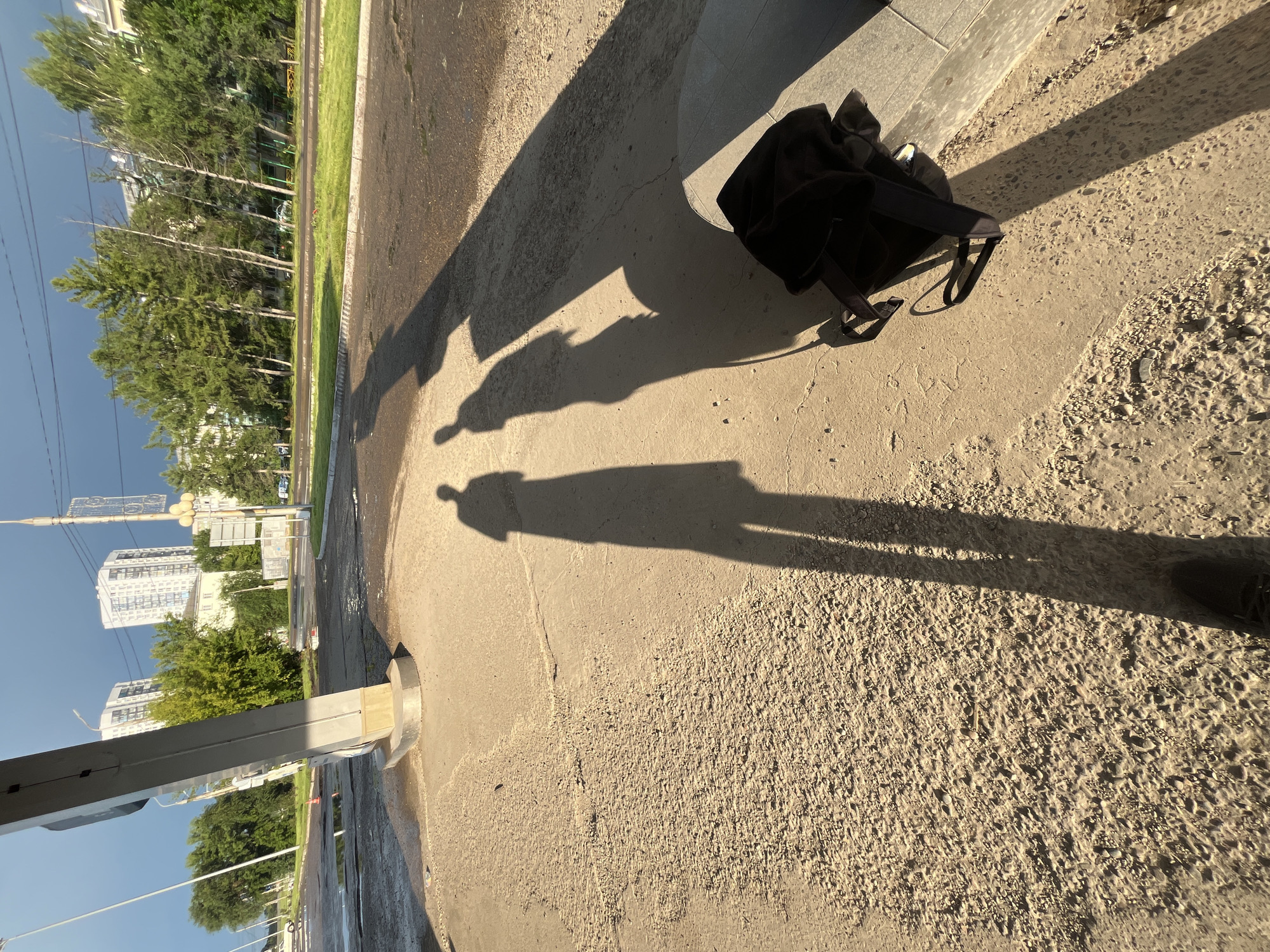 In Krasnoyarsk, Russia, the image captures two elongated shadows of individuals projected onto a textured concrete surface under the golden light of late afternoon. The shadows stretch from left to right, suggesting the subjects are facing away from the sun. A dark backpack rests on the ground to the right, casting a faint shadow. In the background, a large, modern building with numerous windows rises above a line of trees, and a utility pole with various attachments stands tall against a dramatic, dark, and cloudy sky, indicative of an approaching storm. The ground in the foreground is a mix of cracked concrete and loose gravel, with some areas appearing wet, possibly from recent rain. The scene depicts an outdoor urban environment during a transitional weather period.