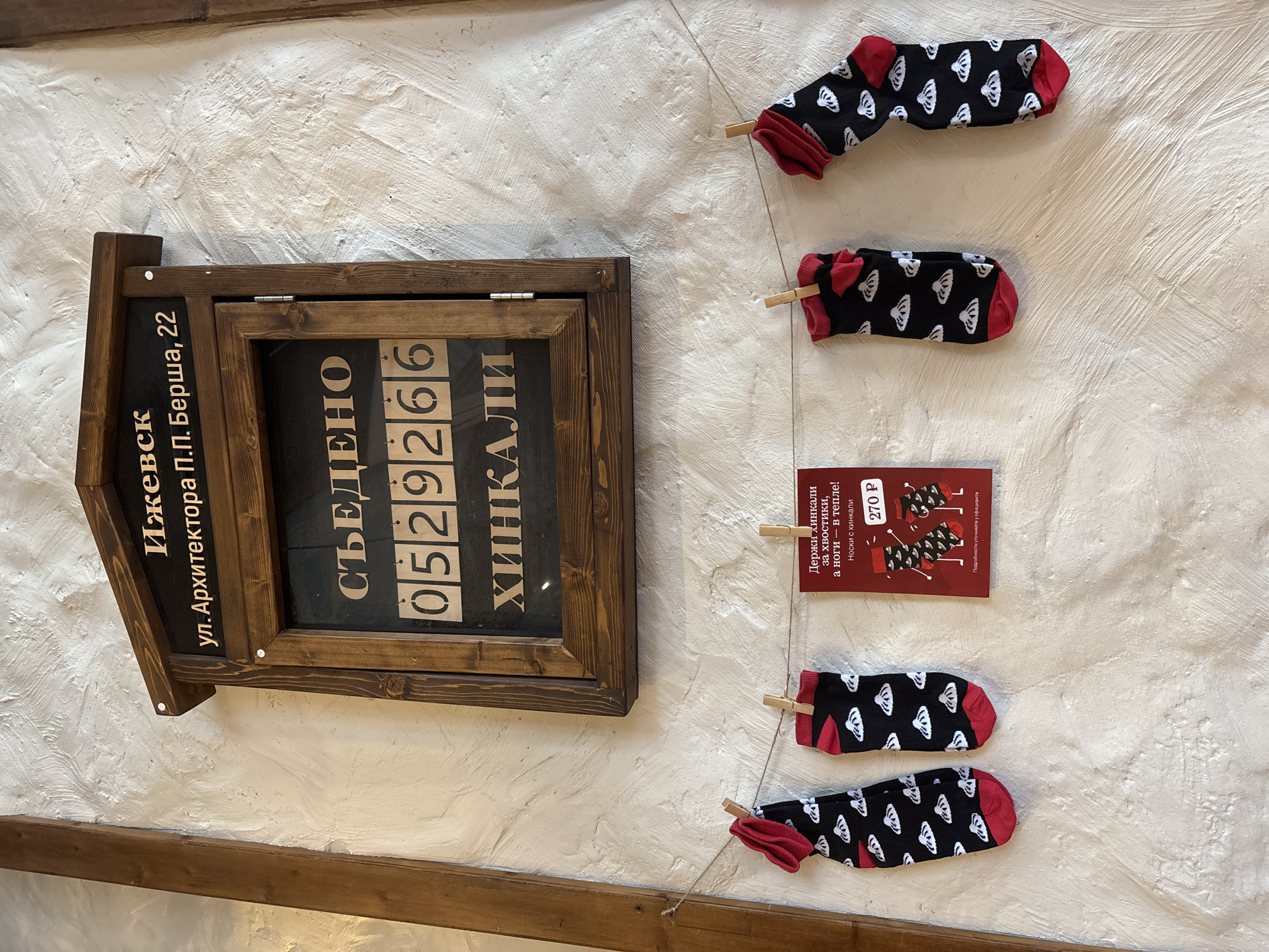 The scene shows a wall display for a Khinkali restaurant in Izhevsk, Russia, specifically located at 22 Arhitektora P.P. Bersha Street. On the wall is a wooden sign showing how many Khinkali have been consumed at the restaurant: 0529266. Beside this sign, black socks with the image of a Khinkali on them hang from clothespins. Beside the socks is a red card displaying a slogan about the Khinkali socks, "Hold Khinkali by their tails, and your feet are warm!", and their price, 270 rubles.