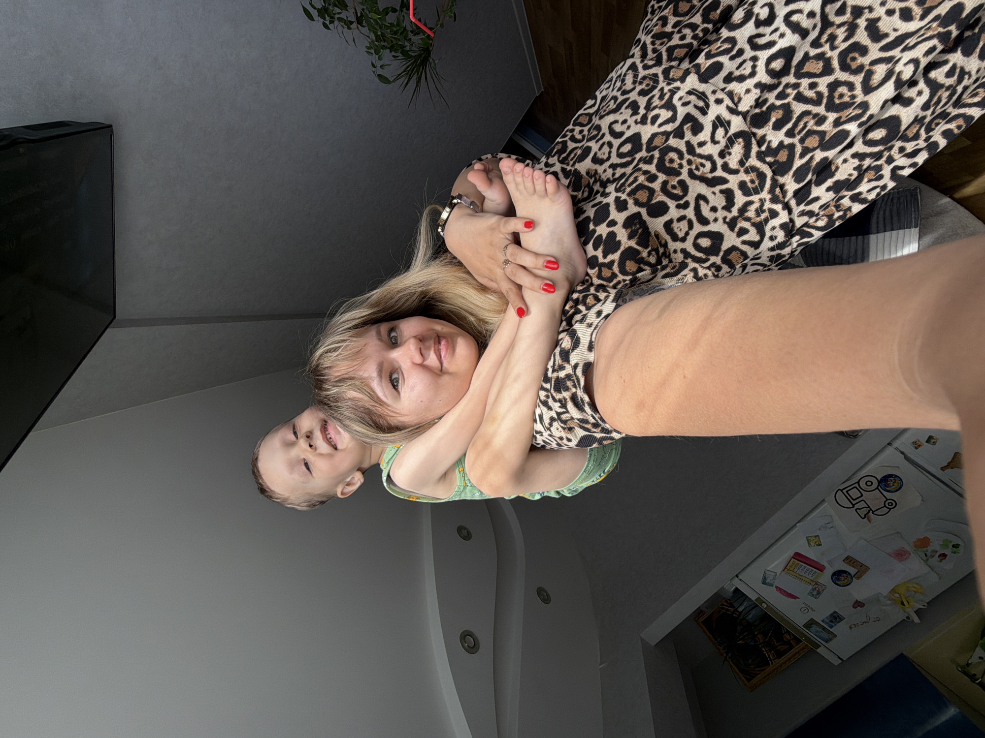 A blonde woman is holding a young child in a room. The woman is wearing a leopard-print shirt and has red nail polish on her fingers. The child is wearing a green tank top. The location is likely in an apartment in Chaikovsky, Russia, evidenced by the presence of household items such as a television, a plant, and a refrigerator. The room has a modern style with gray walls and recessed lighting.