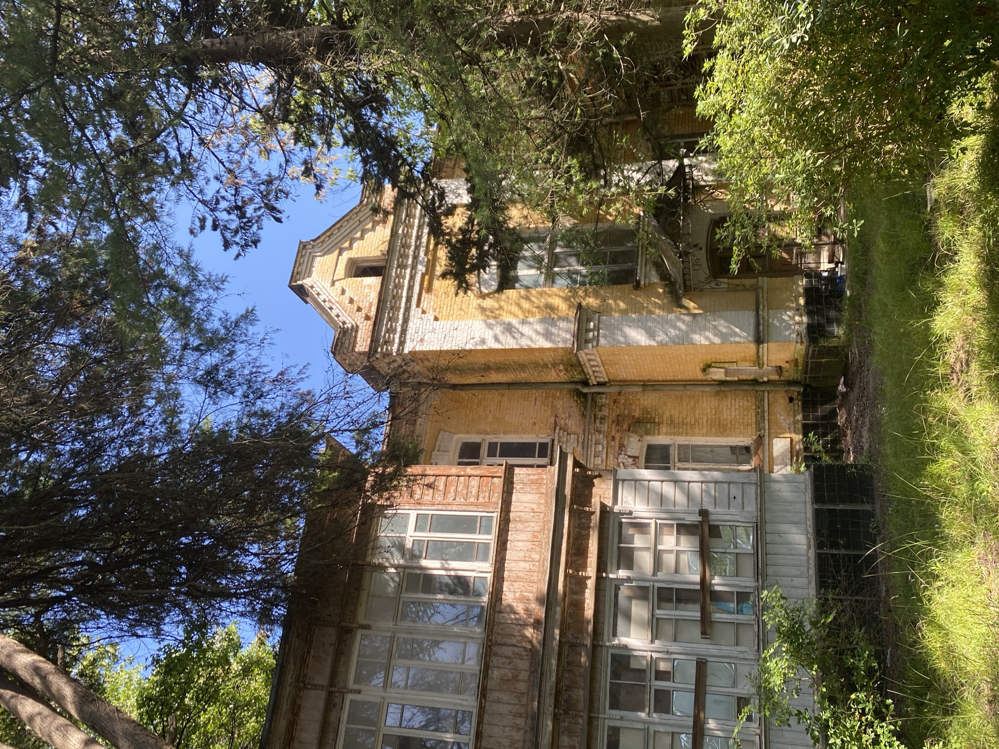 An abandoned, two-story building with yellow brick on the right and weathered wooden siding on the left, is set amidst lush green foliage and tall trees under a clear blue sky. The building's architecture features a pitched roof with a small, arched window on the upper gable, and decorative brickwork. The left side of the building has a wooden balcony with multiple panes of glass windows. The right side of the building has a covered entrance with an ornate metal awning. The foreground is covered in tall, green grass. The image appears to be taken in the city of Sokhumi, Abkhazia, given the architectural style and the surrounding vegetation.