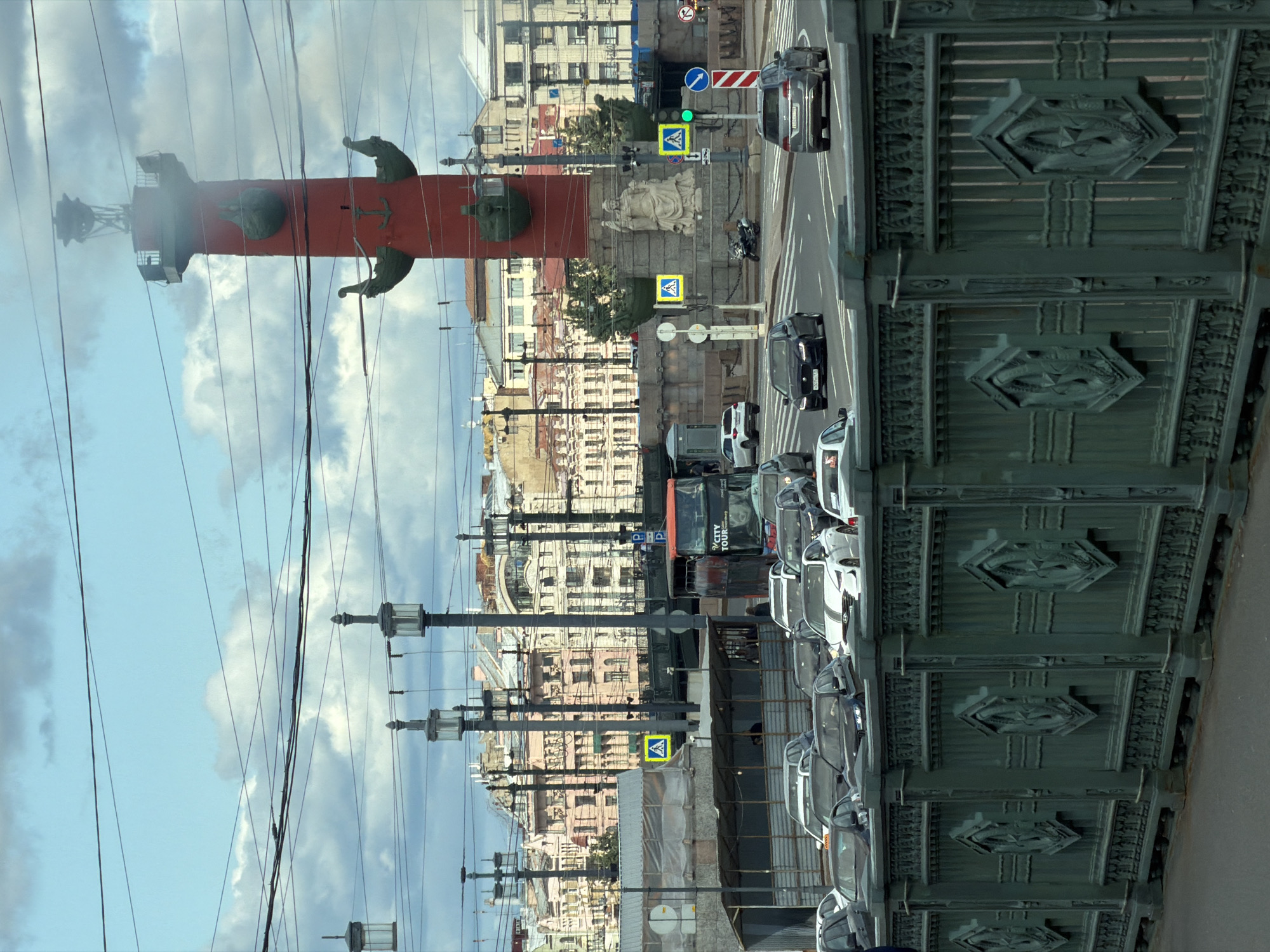 This image captures a street scene in Saint Petersburg, Russia, likely on Vasilyevsky Island. In the foreground, a decorative green metal fence lines a bridge or embankment. Beyond the fence, traffic moves along a street. Numerous cars, including a dark Jaguar and a silver SUV, are visible. To the right, a traffic light displays a green signal, and a directional arrow indicates a lane for turning left.
In the mid-ground, a large, red Rostral Column stands prominently, a significant landmark in the city. The column is topped with a viewing platform and decorated with ship prows and anchors. Behind the column, the neoclassical architecture of Saint Petersburg's buildings is visible, characterized by their light-colored facades and intricate detailing.
Further back, streetlights and overhead wires crisscross the sky, indicating an urban environment. A double-decker tour bus, with "CITY TOUR" branding, is stopped in traffic. The sky is a mix of blue and clouds, suggesting overcast weather. Several pedestrian crossing signs, featuring the universal symbol of a person crossing, are positioned near the street. A statue of a seated figure, possibly historical or allegorical, is located at the base of the Rostral Column. A motorcycle is parked near the statue.