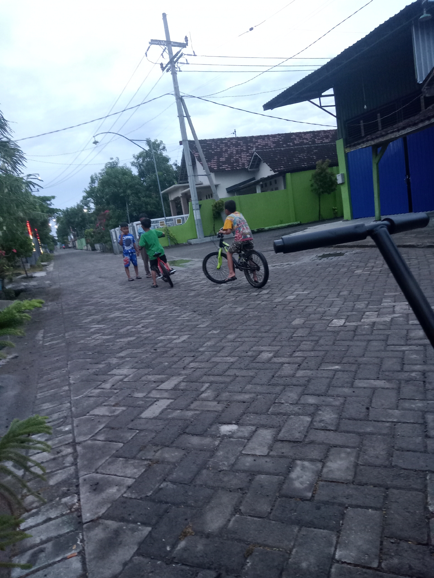 The photograph depicts a residential street in Bungur, Indonesia. The street is paved with dark gray bricks. Two boys are standing near the side of the street, one wearing a blue patterned shirt and shorts, and the other wearing a green shirt. A third boy is riding a bicycle further down the street, wearing a yellow patterned shirt. Houses with green and blue exterior walls are visible in the background, along with power lines and a utility pole. A black object, possibly part of a scooter, is partially visible in the foreground on the right side of the image. The sky is overcast.