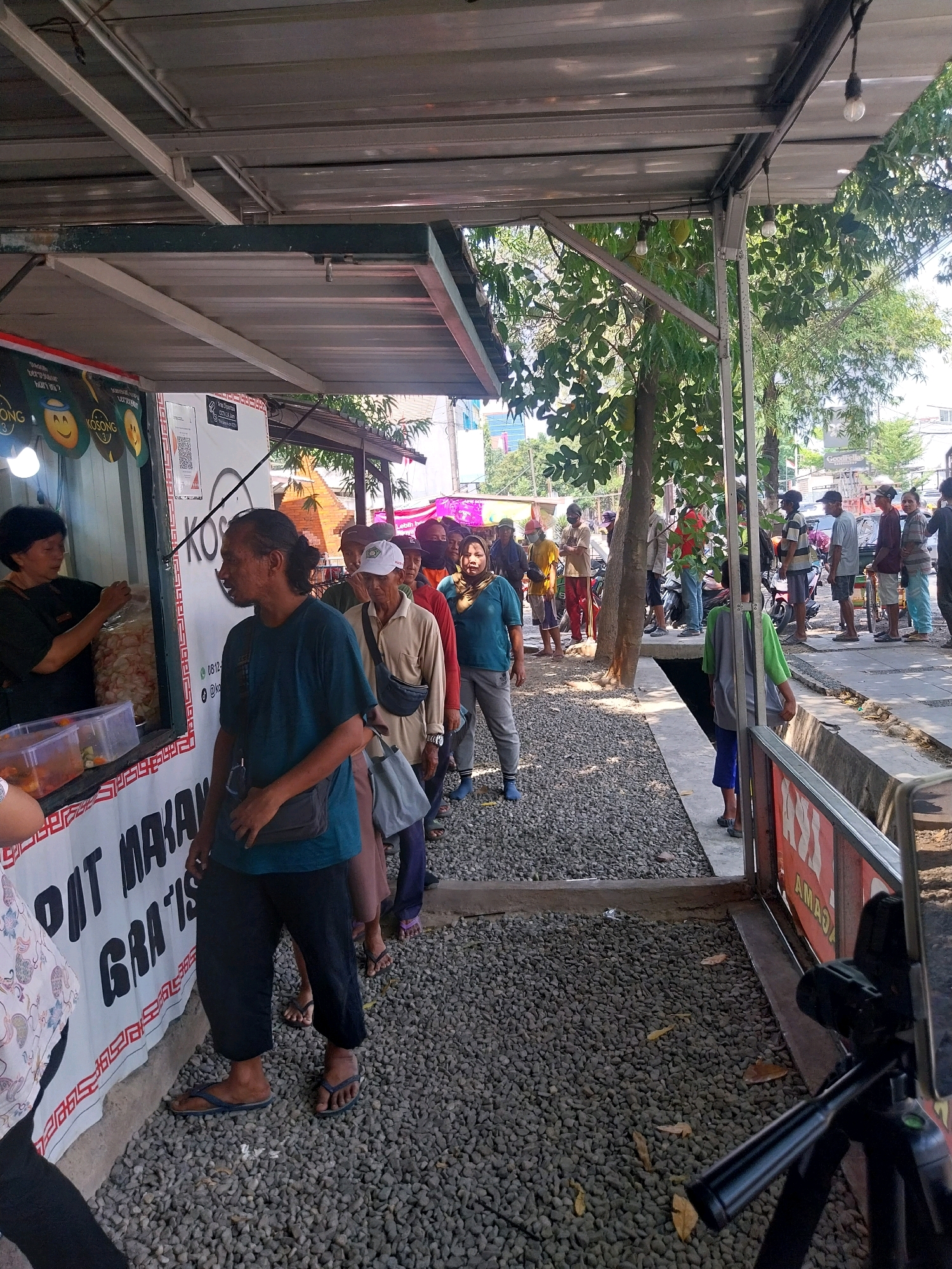 The image depicts an outdoor scene, likely a food stall or market, in Cirebon, Indonesia. A line of people are waiting to purchase food from a stall. The stall has a white banner with red and black text, some of which is legible, including "DAT MAKAN GRATIS". A woman is seen serving food from behind the counter. The ground is covered with gravel, and there are trees and buildings visible in the background. The overall atmosphere appears to be casual and bustling.