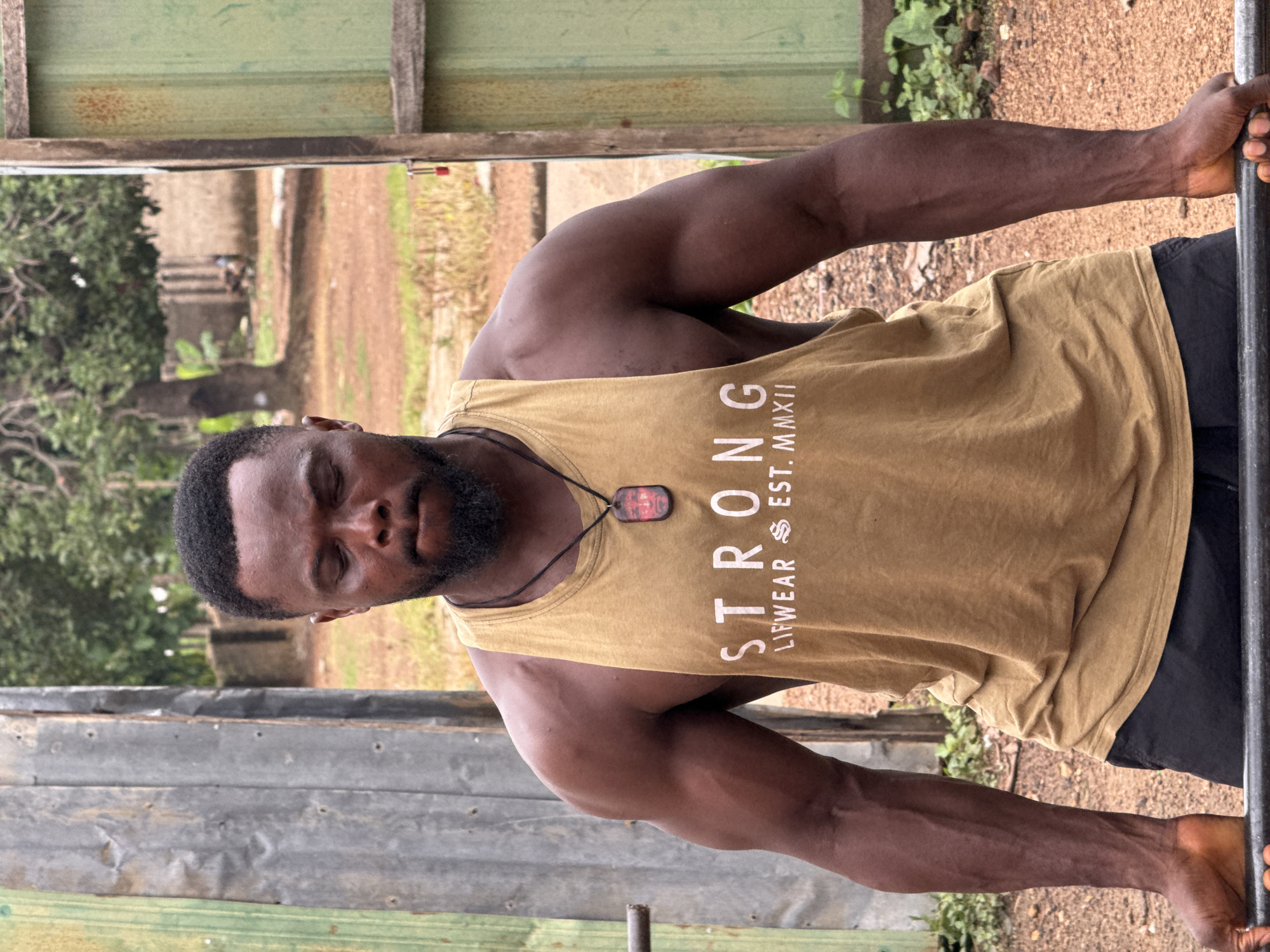 A Black male with a beard, wearing a tan tank top with the word "STRONG" and "LIFWEAR EST. MMXXII" printed on it, and a pendant around his neck, is standing with his eyes closed, gripping a barbell. He is in an outdoor setting with a dirt ground and some greenery. The background shows corrugated metal structures and trees. The location is specified as Akwanga, Nigeria.