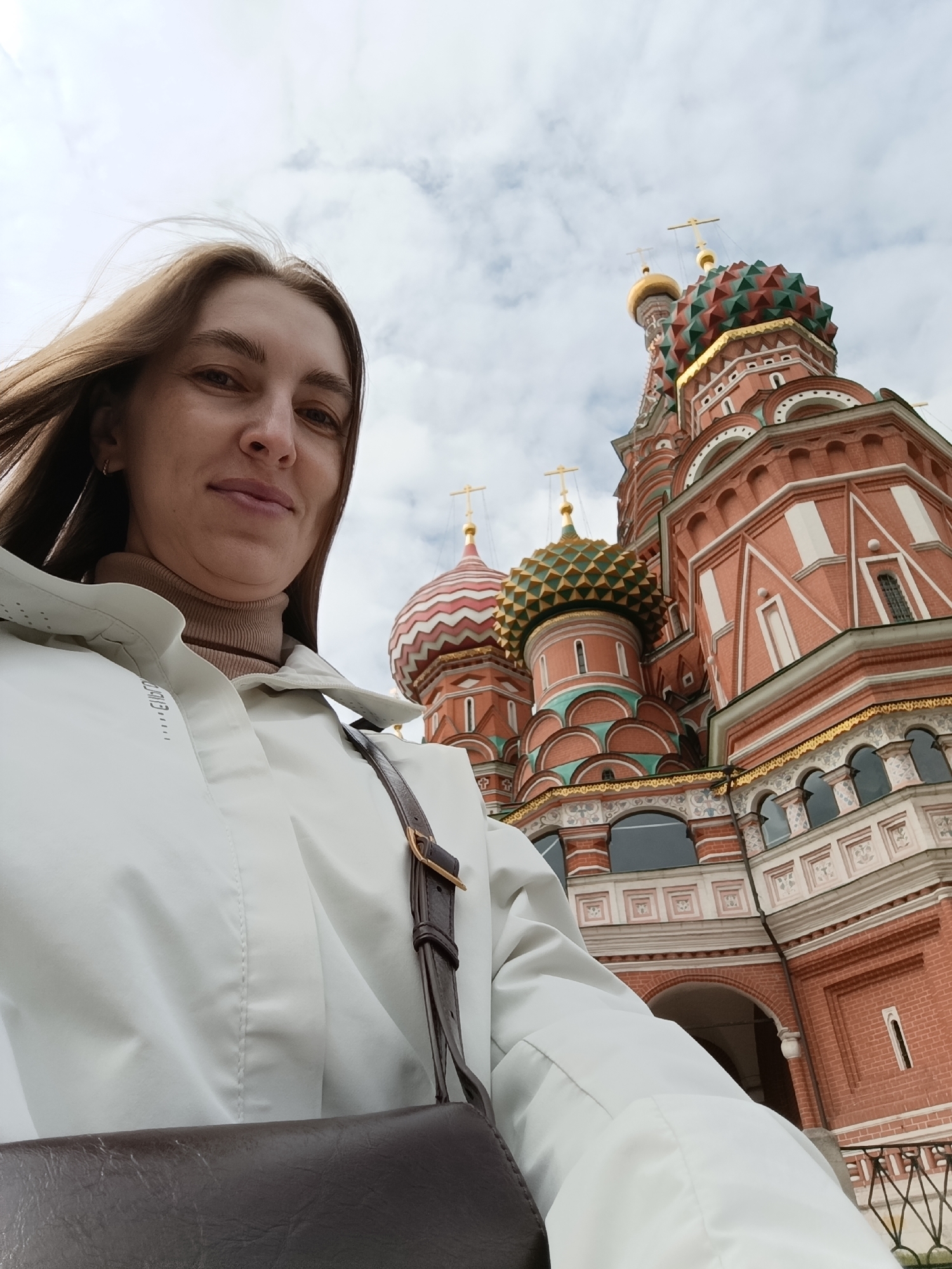 A woman is captured in a close-up selfie, looking directly at the camera with a slight, pleasant smile. She has long, dark blonde or light brown hair, which appears to be gently swept back by a breeze. She is wearing a light-colored (possibly white or very light grey) jacket with a high collar over a beige turtleneck. A dark brown shoulder bag strap is visible across her chest.
Behind the woman, dominating the background, is a clear view of Saint Basil's Cathedral in Moscow, Russia. The cathedral is distinguished by its vibrant, ornate onion domes, which are painted in various patterns of red, green, and gold, and topped with golden spires and crosses. The main structure of the cathedral is made of red brick with intricate white decorative elements and arched windows.
The sky above is overcast with visible clouds, providing soft, diffused lighting for the scene. The time of day appears to be daytime. The woman's expression suggests she is enjoying her visit to the iconic landmark. Faint, indistinct white text or a decorative pattern is visible on the left side of her jacket.