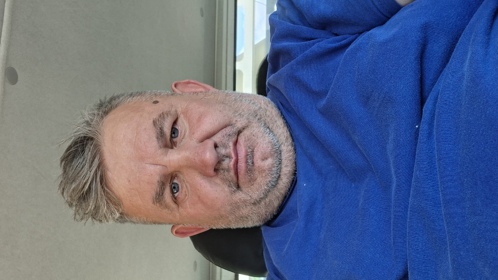 A medium shot, eye-level, selfie-style photograph shows a middle-aged Caucasian man with fair skin, blue eyes, and short, tousled grey hair. He has a salt-and-pepper beard and stubble, and noticeable submental fat. A small, dark mole is visible on his right cheekbone. He is wearing a bright blue t-shirt with a crew neck. The background suggests an indoor setting, possibly a vehicle, with a light-colored ceiling and a dark headrest visible behind him. To the right, a blurred view of the outdoors, with windows and light, indicates it is daytime. The image is taken in Hîncești, Moldova.