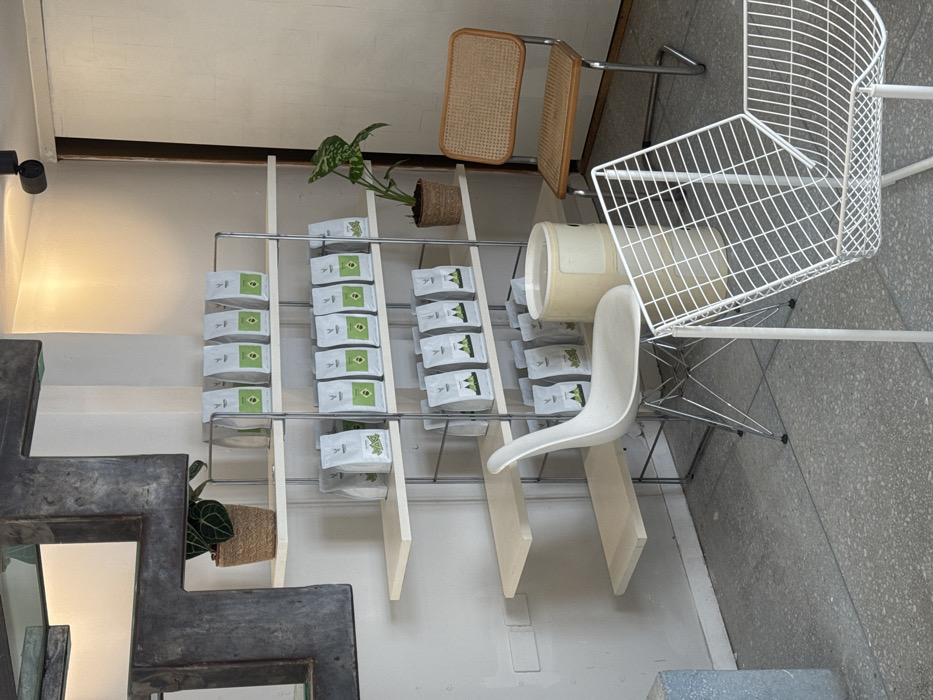 The scene depicts an interior commercial space located in Tel-Aviv, Israel. A prominent metal and light wood shelving unit occupies the center, displaying multiple rows of retail products. The unit features four shelves, each holding approximately eight white pouches with green branding, totaling around 32 packages. Pouches on the bottom two shelves feature a green mountain graphic, while the top two display a simpler green square.
To the far left, a large, dark, irregularly shaped decorative piece, possibly metal or concrete, is mounted on a light-colored wall. Below it, on the top shelf, a small plant in a woven pot is visible. Another similar plant in a woven pot rests on the second shelf from the top, to the right of the packaged products.
The foreground and right side of the image feature various seating options. A white Eames-style rocking chair with a chrome wire base is positioned near the shelving unit. Further right, a brown rattan-backed chair with a chrome frame faces the display. A beige cylindrical object, possibly a planter or receptacle, sits between this chair and the shelving. Partially visible in the immediate foreground, a white metal wireframe chair with an angular design is present. The floor is a light gray concrete or terrazzo. The walls are light-colored, and a black track light fixture is mounted on the ceiling on the right side. No individuals or specific actions are depicted in the static scene.