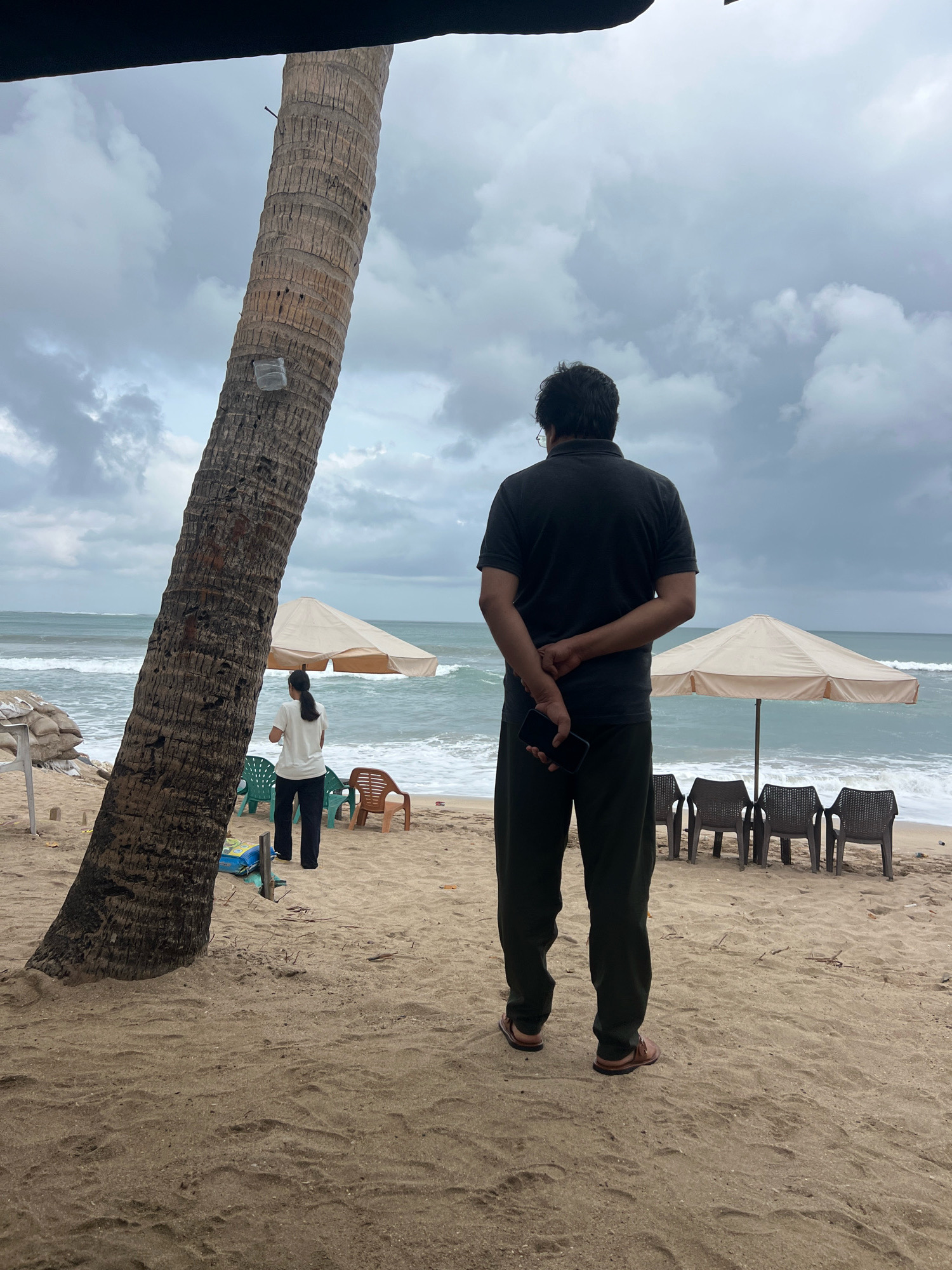 A man stands on a sandy beach, his back to the camera, looking out at the ocean. He is wearing a dark polo shirt and dark pants, with his hands clasped behind his back. In his right hand, he holds a dark smartphone. Behind him, two beach umbrellas are set up, with several empty plastic chairs lined up in front of them. A large palm tree trunk dominates the left side of the frame. Further down the beach, a woman in a white shirt and dark pants is visible, standing near some chairs. The sky is overcast, and the waves are rolling onto the shore. This scene is likely located in Kuta, Indonesia, a popular beach destination known for its surfing and lively atmosphere.