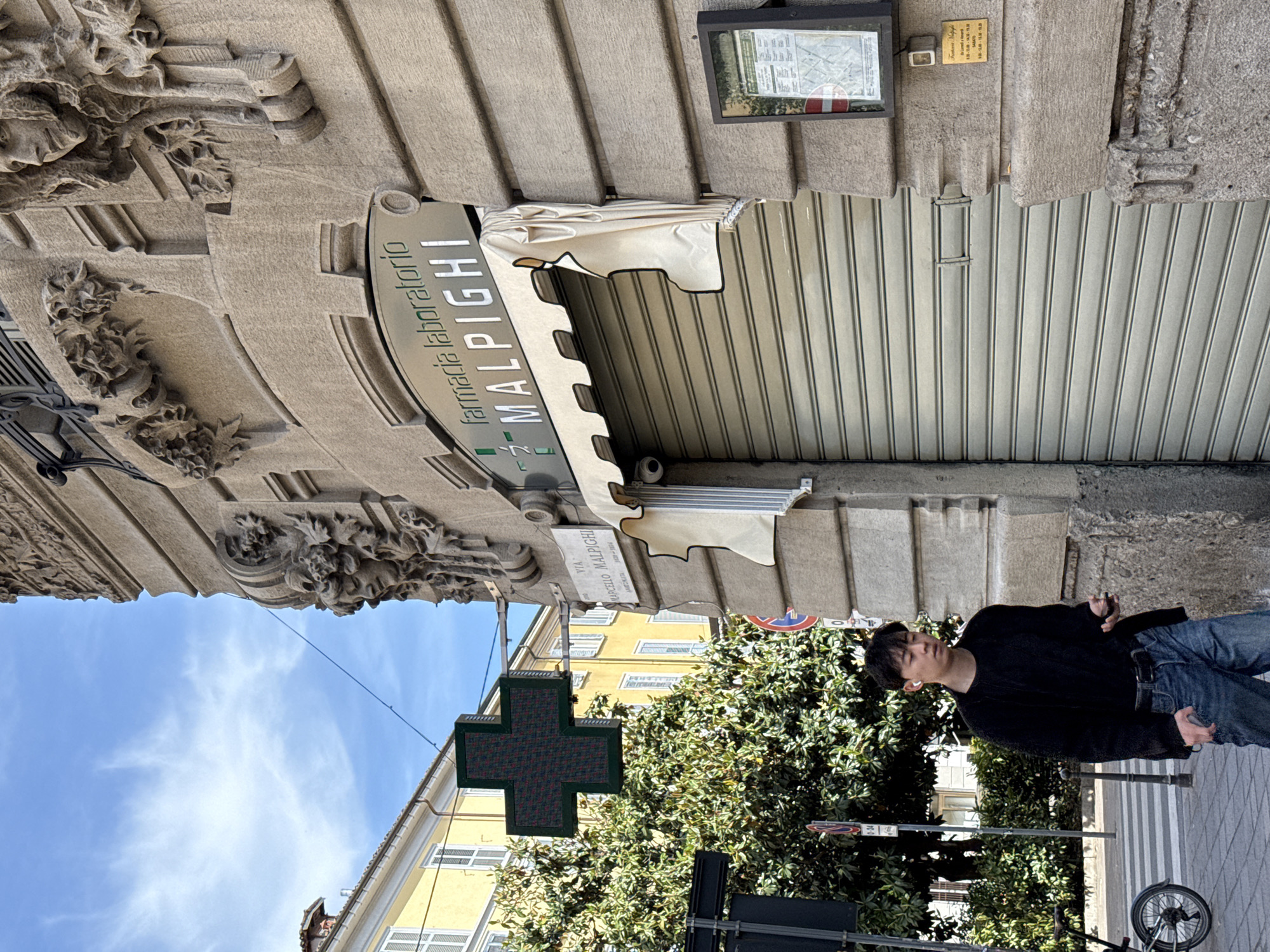 The image shows a street scene in Milan, Italy. The camera is tilted, creating a disorienting effect. A man with dark hair, wearing a black shirt, blue jeans, and white earbuds, is walking on the sidewalk. A bicycle is parked nearby. The sidewalk is paved with light-colored stones. Behind the man are trees and traffic signs. On the left, a pharmacy sign "farmacia laboratorio MALPIGHI" hangs over a building. A digital pharmacy cross is visible further up the street on another building. The architecture suggests an older part of the city. The building facades are predominantly yellow with white window frames. Above the pharmacy, the awning is closed and the security shutter is down. A building plaque reads "Via Marcello Malpighi, Anatomista 1628-1694". The sky is blue with scattered clouds.