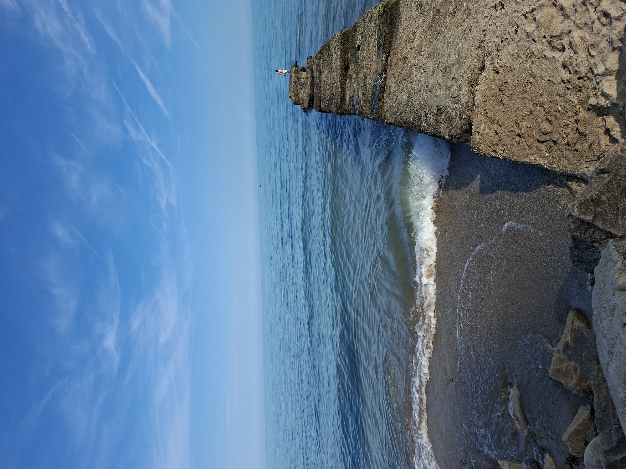 The image displays a coastal scene in Varna, Bulgaria, under a clear, bright blue sky. The sky contains scattered wispy white cirrus clouds and multiple linear contrails. A calm, deep blue body of water, likely the Black Sea, dominates the mid-ground, extending to a faint horizon line. Gentle ripples are visible across the water's surface, and a small, dark, distant object, possibly a boat, is present in the far left-midground.
On the right side of the image, a weathered, multi-segmented concrete pier or breakwater extends from the sandy shore into the sea. The pier's surface is uneven, exhibiting a rough texture with visible signs of age, erosion, and organic growth such as moss or algae. Waves are breaking against the base of the pier, generating white foam and splashes.
At the far end of this pier, a single individual, appearing to be a child or small adult, stands facing away from the camera, looking out towards the open sea. The person is wearing light-colored shorts or swim trunks.
To the left of the pier, a narrow strip of sandy beach is visible, where small waves are breaking and receding. The sand is a light brown-gray color. In the immediate foreground, dark rocks and stones are partially visible on the sandy and wet areas of the shore.