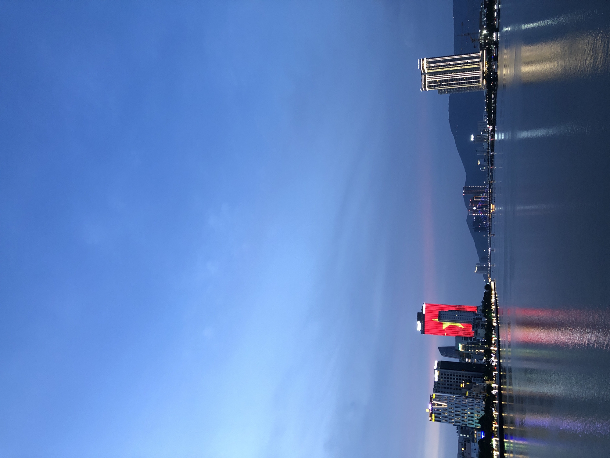 This image captures a twilight scene in Da Nang, Vietnam, featuring a city skyline along a river. In the foreground, the calm river reflects the illuminated buildings on the opposite bank. The most prominent structure is a tall building displaying a large red flag with a yellow star, likely the flag of Vietnam, due to its prominent placement and the location being Da Nang, Vietnam. To the left, several other buildings are lit up with various patterns of lights. To the right, another modern skyscraper stands illuminated with vertical white lights. The background consists of distant hills and a serene, deep blue sky with faint clouds, suggesting the transition from day to night. The overall atmosphere is peaceful and urban. There are no people visible in the image.