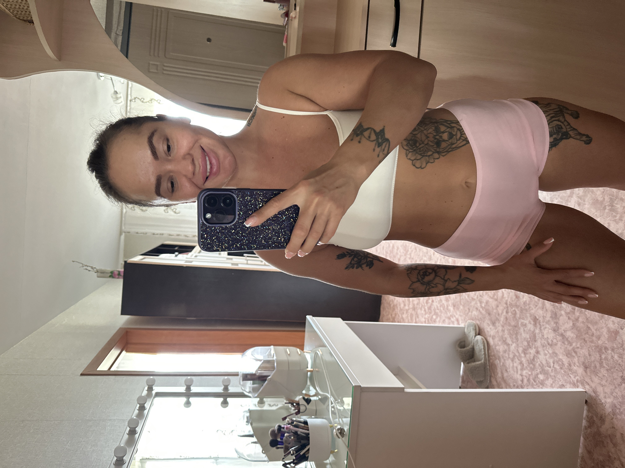 A woman is taking a mirror selfie in a room. She is wearing a white sports bra and pink shorts. She has tattoos on her arm, stomach, and thigh. The room appears to be a bedroom or dressing area, with a vanity table and mirror on the left, and a closet on the right. The floor is carpeted in a light pink color. The vanity has makeup brushes and containers on it. In the background, a door and a window can be seen. The image is taken in Yuzhnouralsk, Russia.