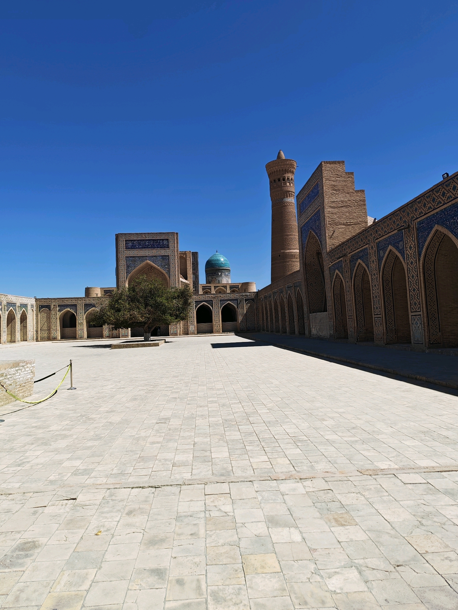 This is an outdoor, eye-level, full shot of the courtyard of the Lyab-i Hauz complex in Bukhara, Uzbekistan. The courtyard is paved with large, light-colored square tiles. In the center of the courtyard, a single, mature tree with a dense canopy of green leaves stands within a raised, brick-bordered planter.
To the left of the courtyard, there is a long, low building with an arched walkway. The arches are decorated with intricate blue and white tile patterns. Beyond this building, and partially obscured by the tree, is a larger structure with an impressive, arched entranceway adorned with similar blue and white tilework.
Dominating the right side of the image is a tall, cylindrical minaret made of brick, with decorative bands and a conical cap. Adjacent to the minaret is another section of the building with a long, arched arcade, also featuring the distinctive blue and white tiling on its facade. A bright turquoise dome is visible above the central building, behind the tree.
The sky is a clear, vibrant blue. A thin yellow rope attached to posts runs across the left side of the courtyard, indicating a boundary. The scene is bathed in sunlight, casting shadows across the courtyard and highlighting the architectural details of the complex. There are no people visible in the image.