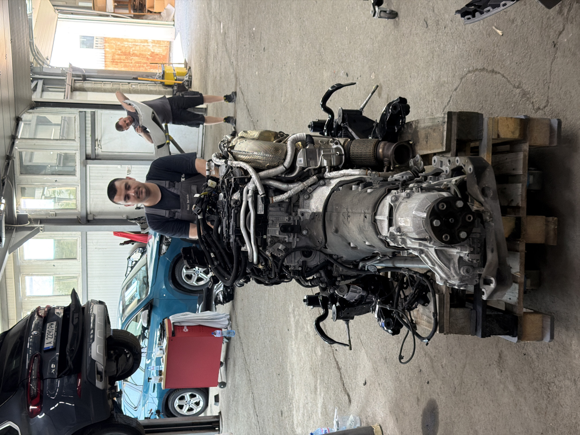 The image shows a man in work overalls standing in front of a large vehicle engine resting on a wooden pallet. The engine is intricate with various wires, hoses, and metal components. In the background, another man is seen holding up a car part. Two vehicles are partially visible behind the engine. One is a dark SUV, the other a light blue car. The setting appears to be a garage or workshop. A red toolbox is visible near the blue car. The license plate "A 2290 PM" is visible on the dark SUV. It is likely the city of Burgas, Bulgaria because a water bottle with Bulgarian Cyrillic characters appears to be placed on the floor.