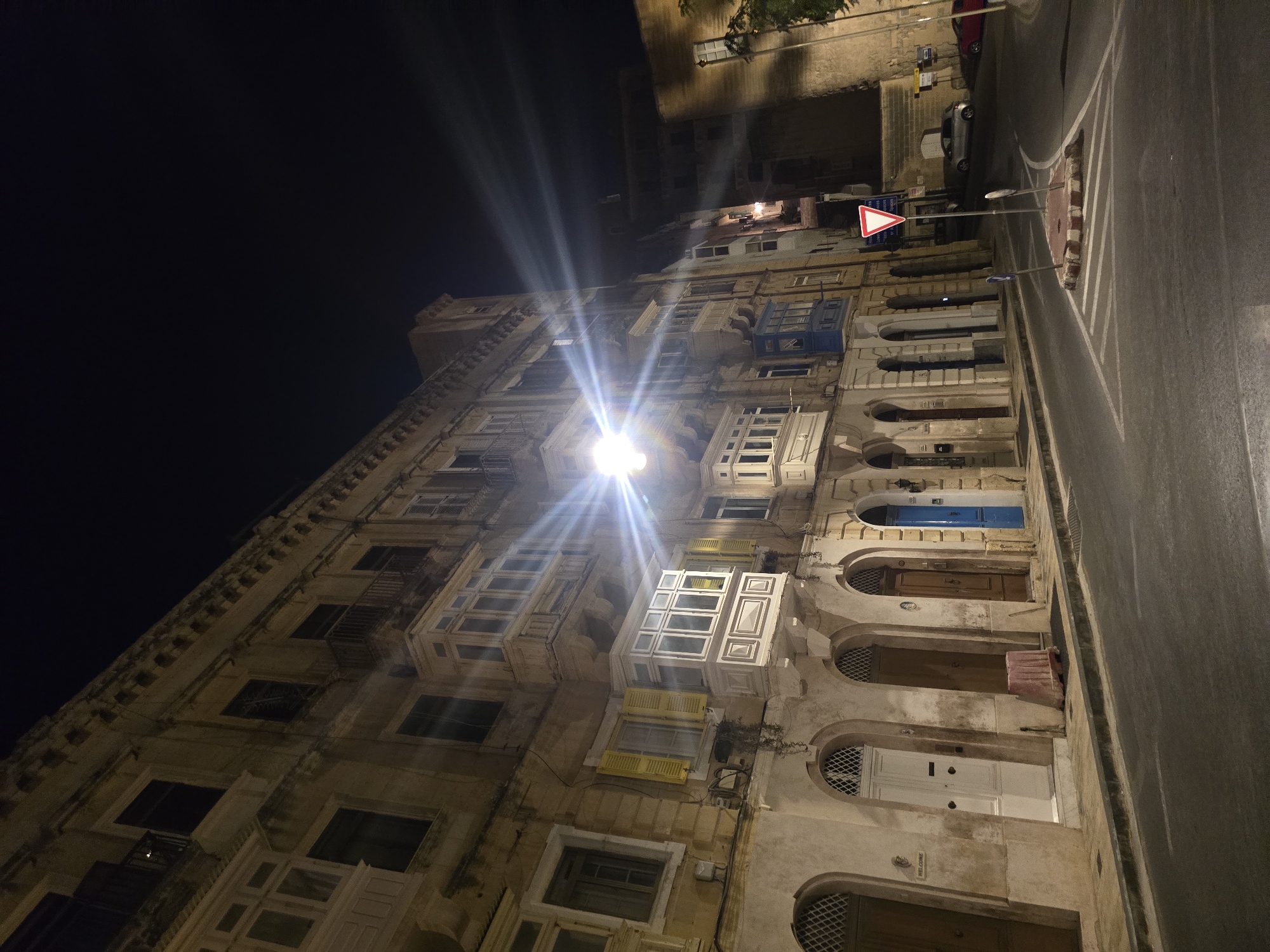 A street scene in Valletta, Malta, at night. The image captures a section of a building facade with traditional Maltese enclosed wooden balconies, known as "gallariji." The architecture features stone construction with arched doorways and windows. A street lamp casts a bright light, creating lens flare and illuminating the asphalt road and sidewalk. Several doors are visible along the ground floor, some painted in muted colors, others in a distinctive blue. A traffic island with a red and white striped border and a yield sign is in the foreground, positioned on the road. A few cars are parked on the street. The overall atmosphere is urban and nocturnal, with the dark sky contrasting with the illuminated building and street.