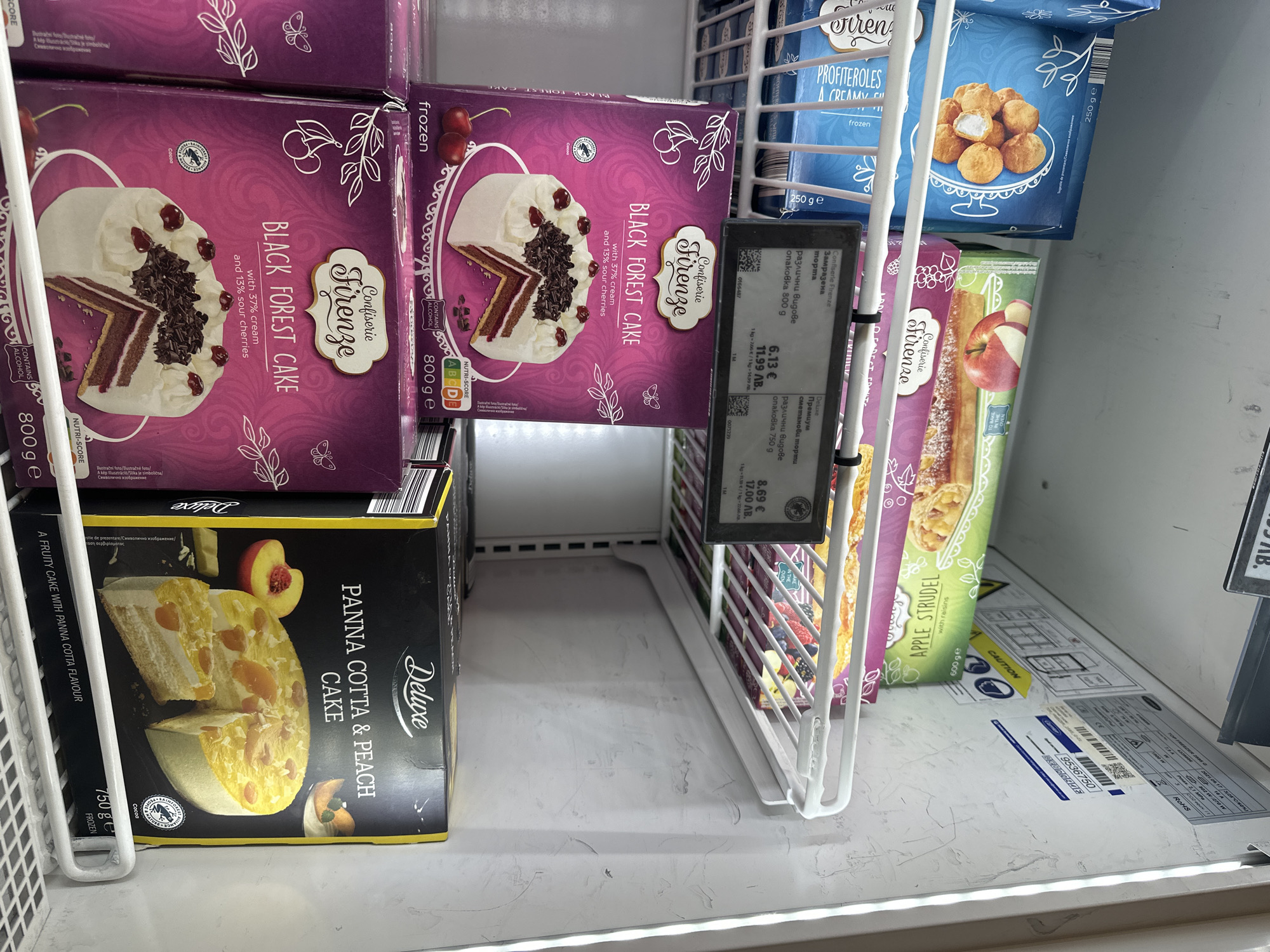 The image depicts a refrigerated section of a store, likely a supermarket, in Sofia, Bulgaria. The refrigerated shelves display various frozen desserts, including "Black Forest Cake" (labeled in both English and Bulgarian), "Panna Cotta & Peach Cake", "Apple Strudel", and "Profiteroles". Price tags in Bulgarian currency (Lev) are visible. The "Black Forest Cake" boxes show a Nutri-Score and alcohol content. A warning sign is visible on the shelf.