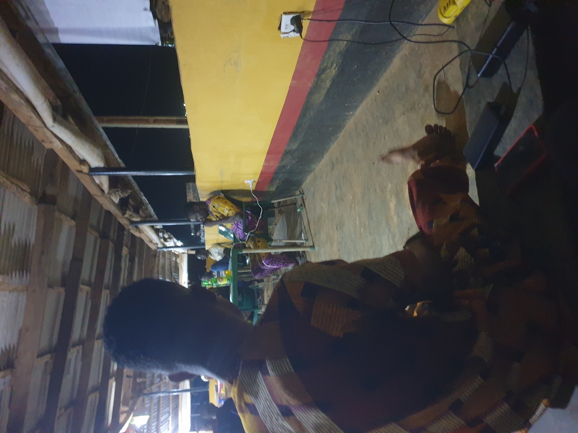 An outdoor, open-air gathering space in Anilelerin, Nigeria, is depicted. The scene appears to be an informal eating or meeting area with a corrugated metal roof supported by wooden beams. The back wall of the structure is painted yellow with a red and black stripe running horizontally along its base. Several people are present, seated at tables and on chairs, with some appearing to be eating or drinking. In the foreground, a person wearing a colorful patterned shirt is seated, facing away from the camera. Electronic devices, including power banks and cables, are visible on the ground near the foreground subject, suggesting charging activity. The lighting suggests it is either late evening or night.