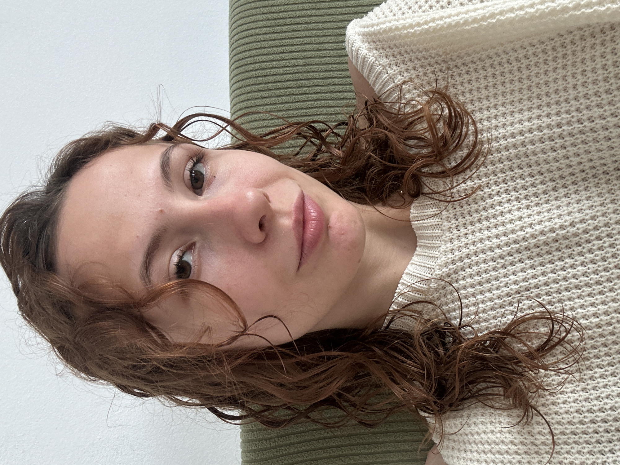 A close-up, canted-angle selfie captures a woman with brown, wavy hair. She is lying on an olive green, horizontally-ribbed surface. She is wearing a textured, off-white knit sweater. Her skin is fair, and she has brown eyes and pink lips. The background consists of a white wall behind her head. No contextual elements indicate the location as Novi Sad, Serbia.