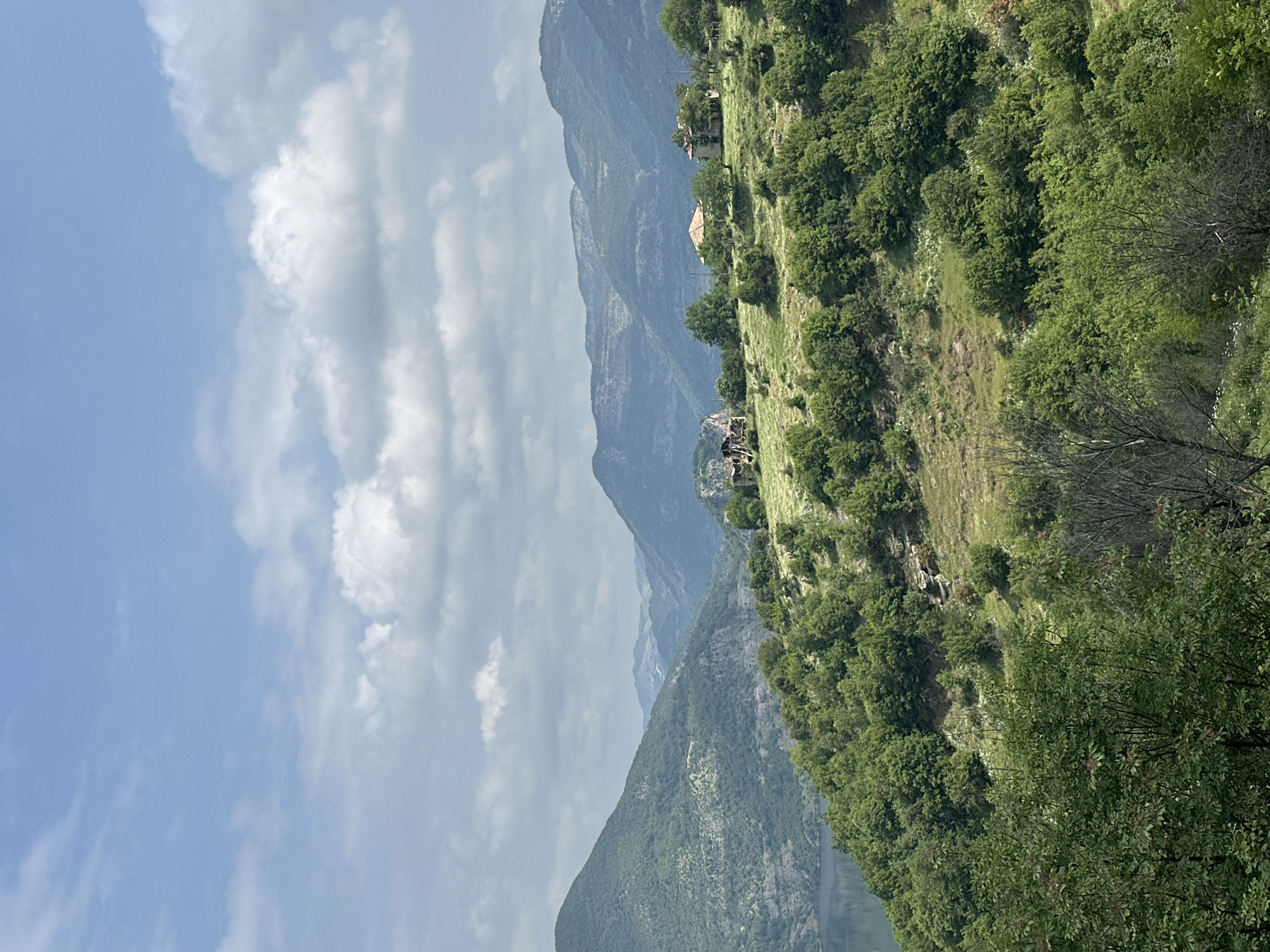 A serene mountainous landscape in Star Chitak, Bulgaria, is captured under a partly cloudy sky during what appears to be daytime, likely late morning or early afternoon. The image is composed of successive layers of natural features, creating a sense of depth and vastness.

In the foreground and midground, a steep hillside slopes from the right, covered in dense, vibrant green foliage. Numerous trees with full canopies dominate this area, interspersed with patches of lighter green grass and some rocky outcrops. Small, isolated structures, possibly houses with light-colored walls and reddish-brown roofs, are nestled amongst the trees on this hillside, indicating human presence within the natural environment.

Beyond this immediate green slope, layers of larger mountains recede into the distance. These mountains exhibit a cooler, bluer hue due to atmospheric perspective, becoming progressively lighter and less distinct. Their ridgelines create a dramatic, undulating silhouette against the horizon. A deep valley or gorge appears to run between these mountain ranges, though its bottom is mostly obscured.

The sky occupies the upper left portion of the image, displaying a mix of clear blue patches and soft, white cumulus clouds. The clouds vary in density, with some appearing bright and voluminous, while others are more diffuse.

There is no discernible activity taking place, as it is a static landscape photograph. No visible text is present in the media file. The overall impression is one of natural beauty, tranquility, and the majestic scale of the Balkan mountain scenery.