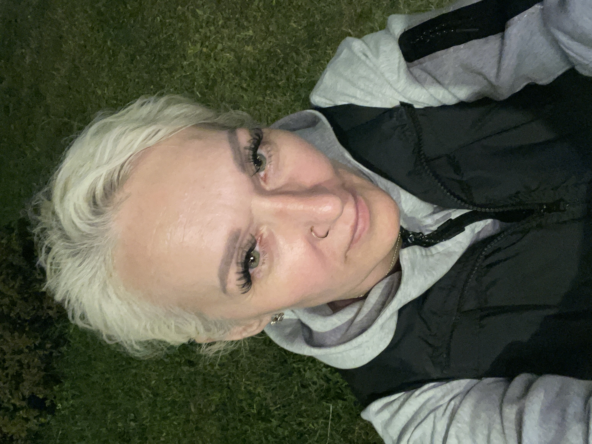 A medium shot, high-angle selfie features a fair-skinned Caucasian woman with short, white, layered hair. She is looking directly at the camera with green eyes and is wearing dramatic false eyelashes. She has a nose ring and a small earring in her left ear. She is wearing a light gray hooded sweatshirt with a black vest zipped over it. The background consists of dark green grass and some foliage, suggesting an outdoor setting at dusk or night, as the lighting appears artificial. No specific location within Saint Petersburg is identifiable from the image.