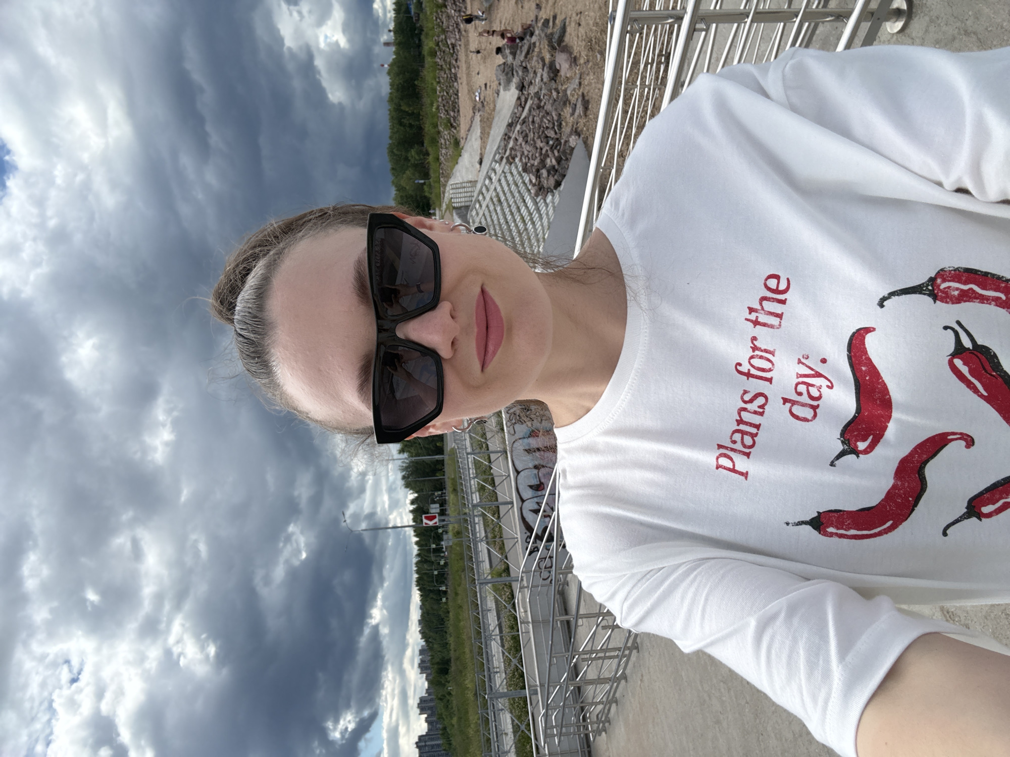 In this selfie, a woman wearing sunglasses and a white long-sleeved shirt with red chili pepper graphics is the main subject. The shirt has text that reads "Plans for the day." She is outdoors with a cloudy sky overhead, suggesting it might be a partly overcast day. The background shows a wooded area, some buildings in the distance, and a metal railing. There are also some rocks and what appears to be a concrete structure in the mid-ground. People can be seen in the distance near the rocks. The scene appears to be a public area, possibly a park or waterfront, given the railing and the open space. The woman's expression is neutral, and the overall mood of the image is casual and observational. The location context points to Saint Petersburg, Russia, which could imply the architectural style of the distant buildings or the type of vegetation might be indicative of this region.