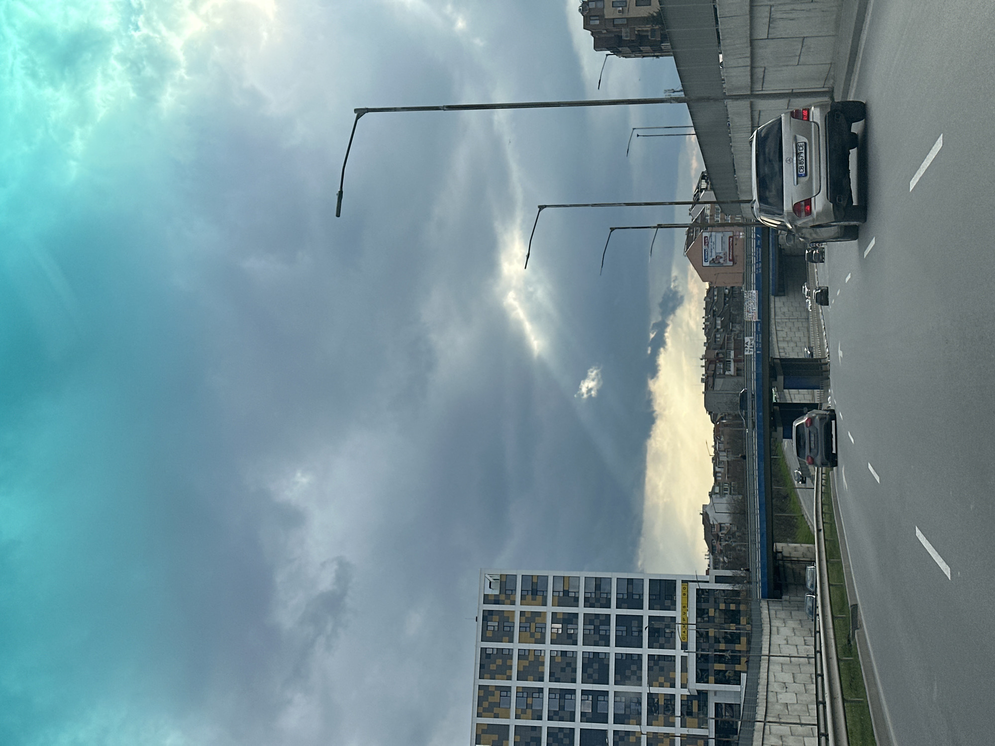 The image depicts an urban landscape, seemingly captured from a moving vehicle on a highway in Varna, Bulgaria. The perspective is tilted, showing a cloudy sky with rays of sunlight breaking through. Adjacent to the highway is a modern building with a grid-like facade, possibly an office or residential complex. A row of streetlights lines the highway. Several cars are visible on the road, including a silver SUV with a license plate number "CB 6579 CE". In the background, there is a cluster of older buildings and apartments, indicative of a more established part of the city. The sky, dominant in the image, transitions from a darker, overcast color to a brighter, light-blue hue on the upper-left portion.