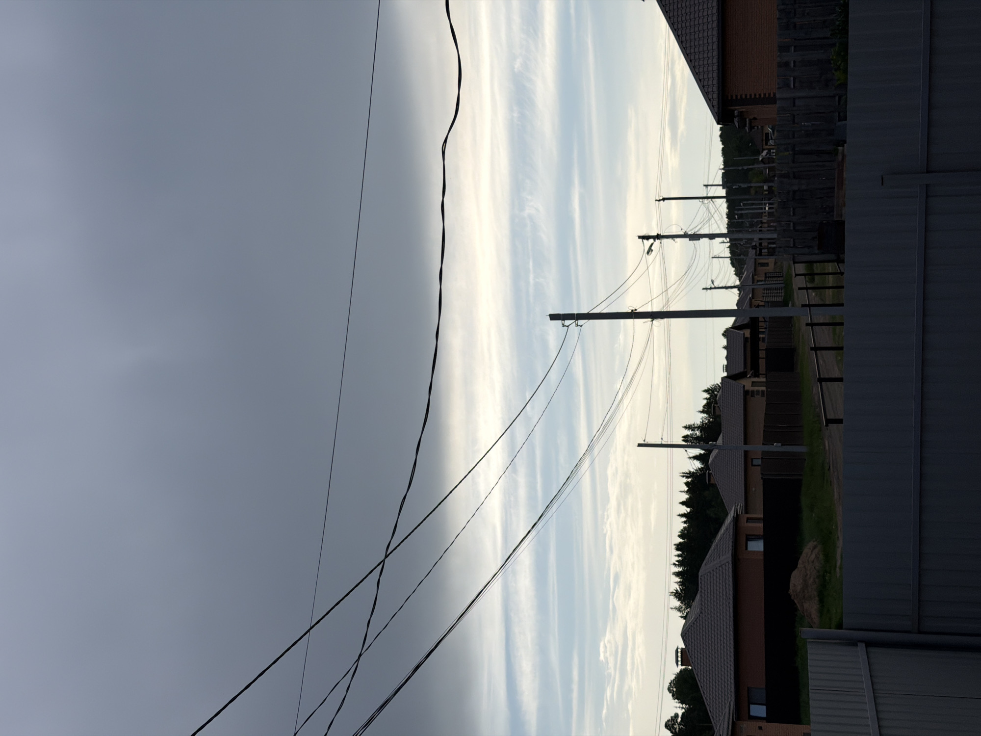The image depicts a residential area in Chaikovsky, Russia. The sky is divided between a dark, overcast area on the left and a brighter, cloud-streaked area on the right. Power lines run across the sky. Houses with brown roofs and brick exteriors are visible, separated by fences. A row of utility poles lines the street, connecting to the houses. The foreground includes a gray metal fence.