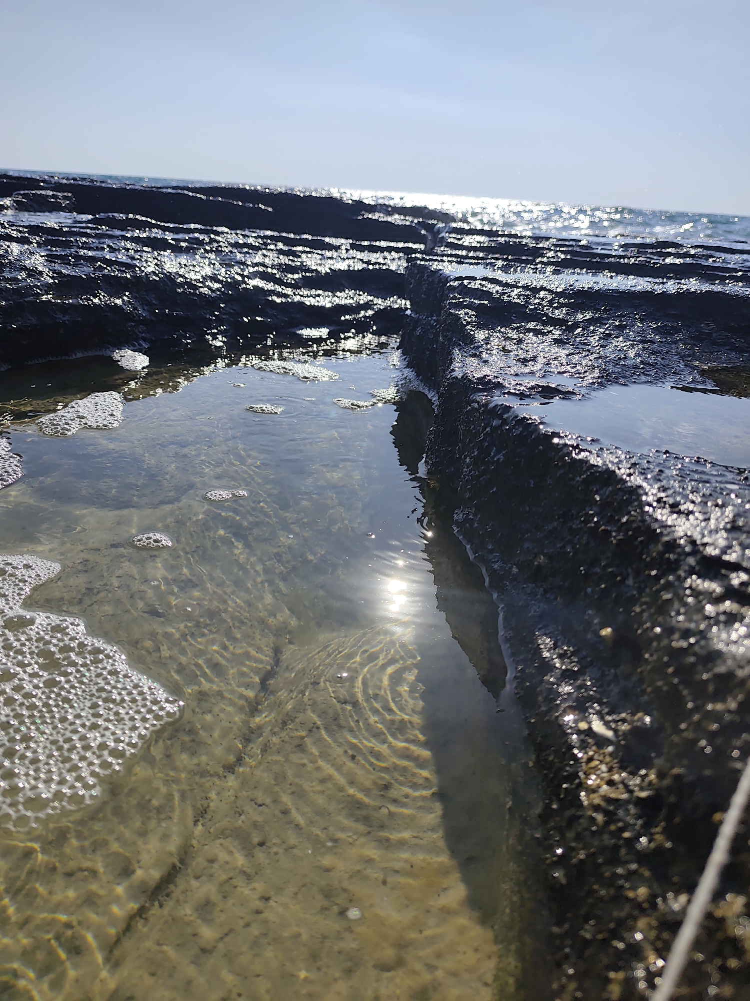 The image depicts a close-up view of tidal pools on a rocky shoreline, likely in Saiyn, Kazakhstan. The sun is shining brightly, creating a sparkling effect on the surface of the sea and reflecting in the shallow water of the pools. The sky is a clear, pale blue, indicating fair weather. The rocks are dark and wet, with some appearing to be volcanic in origin, forming natural channels and basins that hold the water. In the foreground, a large tidal pool is visible, filled with clear water that reveals the sandy and rocky bottom. The water in the pool has a gentle ripple, and there are clusters of small white bubbles along the edge, suggesting recent wave activity or aeration. The sunlight penetrates the water, illuminating the patterns on the seabed and creating a shimmering effect. The overall impression is one of a serene and natural coastal environment bathed in sunlight. There is no visible text in the image.