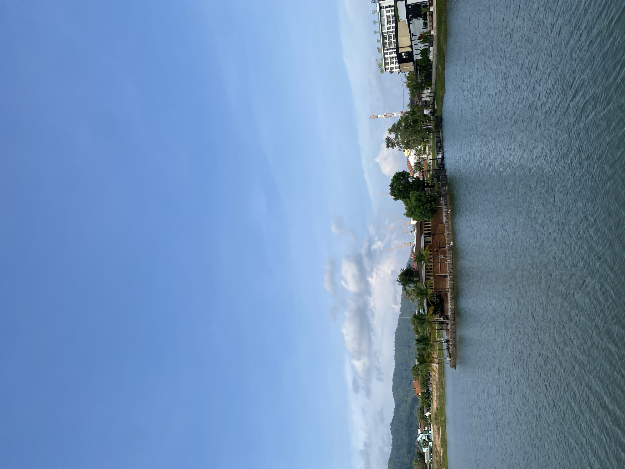 The image presents a serene daytime view of a waterfront area in Choeng Thale, Thailand, viewed from across a body of water. The scene is dominated by a wide expanse of grayish-blue water in the foreground and middle ground, exhibiting subtle ripples on its surface under a bright, clear blue sky with scattered white clouds.
Along the distant shoreline, a diverse landscape unfolds. On the left, a series of modern, multi-story buildings with white facades and dark windows, possibly hotels or residential complexes, stand interspersed with lush green trees and palm foliage. Further along the shore, smaller buildings with red-tiled roofs are visible, nestled amidst dense greenery. Jetties or wooden structures extend from the land into the water at various points.
A prominent white and orange striped tower, resembling a communication tower, rises above the buildings. In the background, a range of verdant mountains creates a natural backdrop, and faint outlines of what appear to be construction cranes can be seen against the sky above them.
The time of day appears to be sunny, likely late morning or early afternoon, with good visibility. The weather is fair, with blue skies and light cloud cover. No individuals or specific activities are discernible within the frame. There is no visible text. The scene depicts a harmonious blend of natural beauty and development in a coastal or lakeside setting.