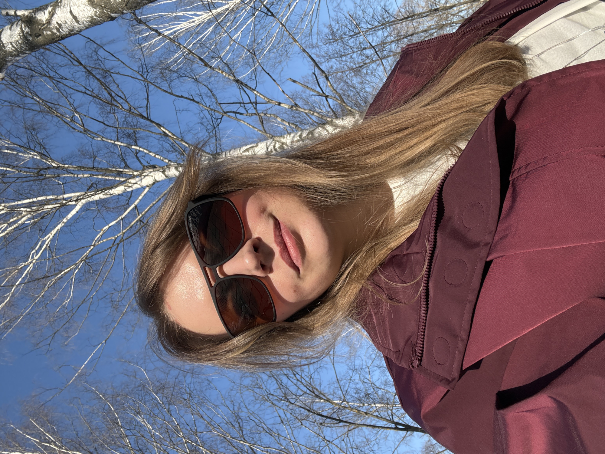The image shows a person wearing sunglasses and a maroon jacket, positioned against a backdrop of bare birch trees and a blue sky. The perspective is angled upwards, suggesting the photo was taken from a low vantage point. The setting is Izhevsk, Russia, recognizable by the characteristic birch trees common in the region. The person's face is partially visible, and the focus is on the upper part of their body and the surrounding natural environment.