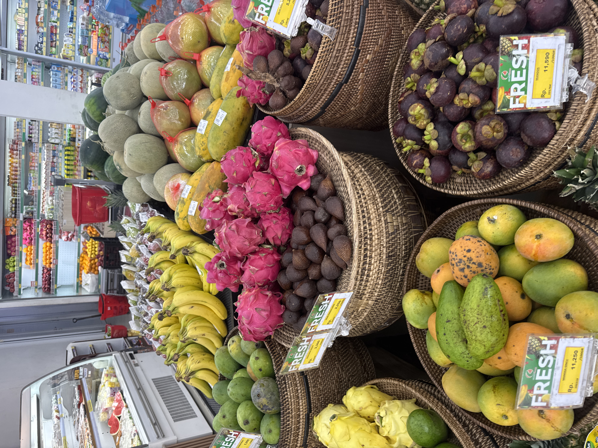 This is a medium-shot, eye-level photograph of a fruit display at a grocery store in Kuta Selatan, Indonesia. The focus is on a variety of fresh fruits arranged in woven baskets. In the foreground, a basket of red dragon fruit is prominent, with some snake fruit in the adjacent basket. To the left, there are green mangoes and some yellow fruits. In the middle ground, a large pile of watermelons and cantaloupes are visible, along with papayas bagged in netting. Behind the fruit display, shelves stocked with various goods are visible, including bottles of liquid and packaged items. The overall lighting suggests an indoor environment, likely a supermarket. No people are visible in the frame.