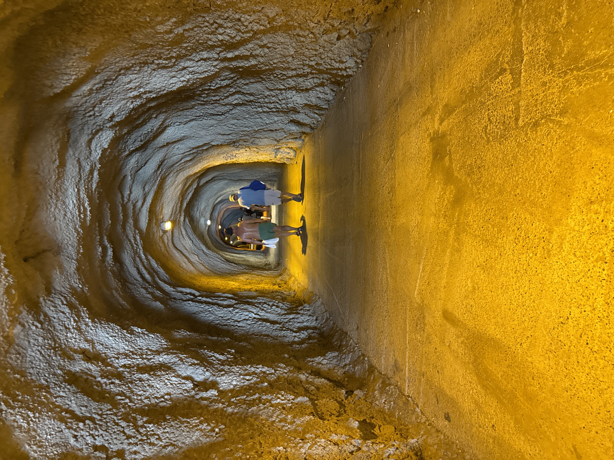 Two individuals are walking away from the viewer down a man-made tunnel with rough, textured walls and a lit path. The tunnel is illuminated by overhead lights, casting a warm, yellow glow. The individual in the foreground is wearing green shorts and is shirtless, carrying a white bag. The individual behind them is wearing a blue shirt and white shorts, and is carrying a blue bag. The tunnel appears to be a pathway through rock, possibly connecting different parts of a coastal area. This tunnel is located in Rafailovići, Montenegro.