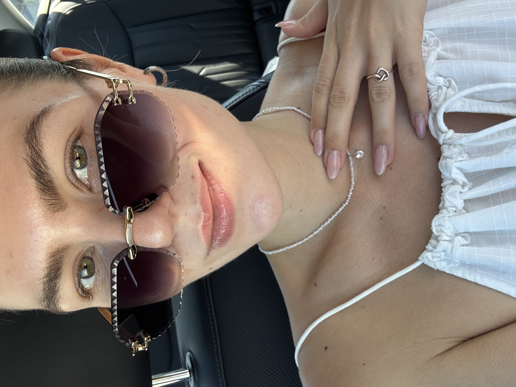 The image is a close-up, angled selfie of a person inside a car. The person has light skin, and their face is partially obscured by large sunglasses with a gold frame. They have green eyes and glossy lips. The person is wearing a thin silver necklace and a ring shaped like a knot on their finger. They are wearing a white top with gathered detailing around the neckline. The interior of the car is visible, with black leather seats.