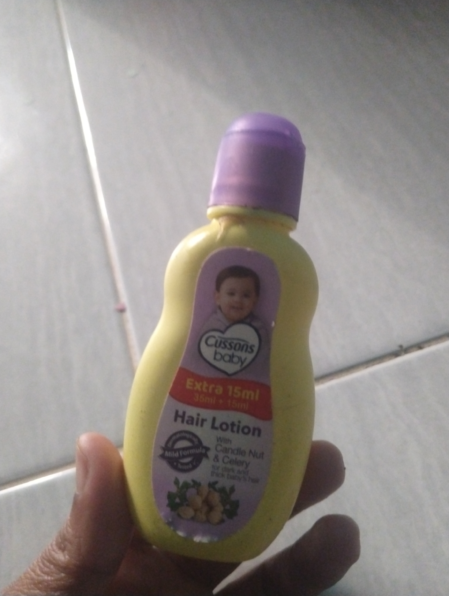 In Wanasaba Daya, Indonesia, a close-up, low-angle shot captures a yellow bottle of Cussons Baby Hair Lotion held by a person's hand. The bottle has a purple cap and a label featuring a baby's face, the text "Cussons Baby," "Extra 15ml," and "Hair Lotion." Ingredients like "Candle Nut & Celery" are also mentioned on the label, along with "Mild Formula." The background consists of a tiled floor, with a prominent vertical line suggesting a grout line or a division between tiles. The lighting appears to be ambient, with some glare on the bottle and floor. The overall scene is a simple, everyday moment, likely in a home environment.