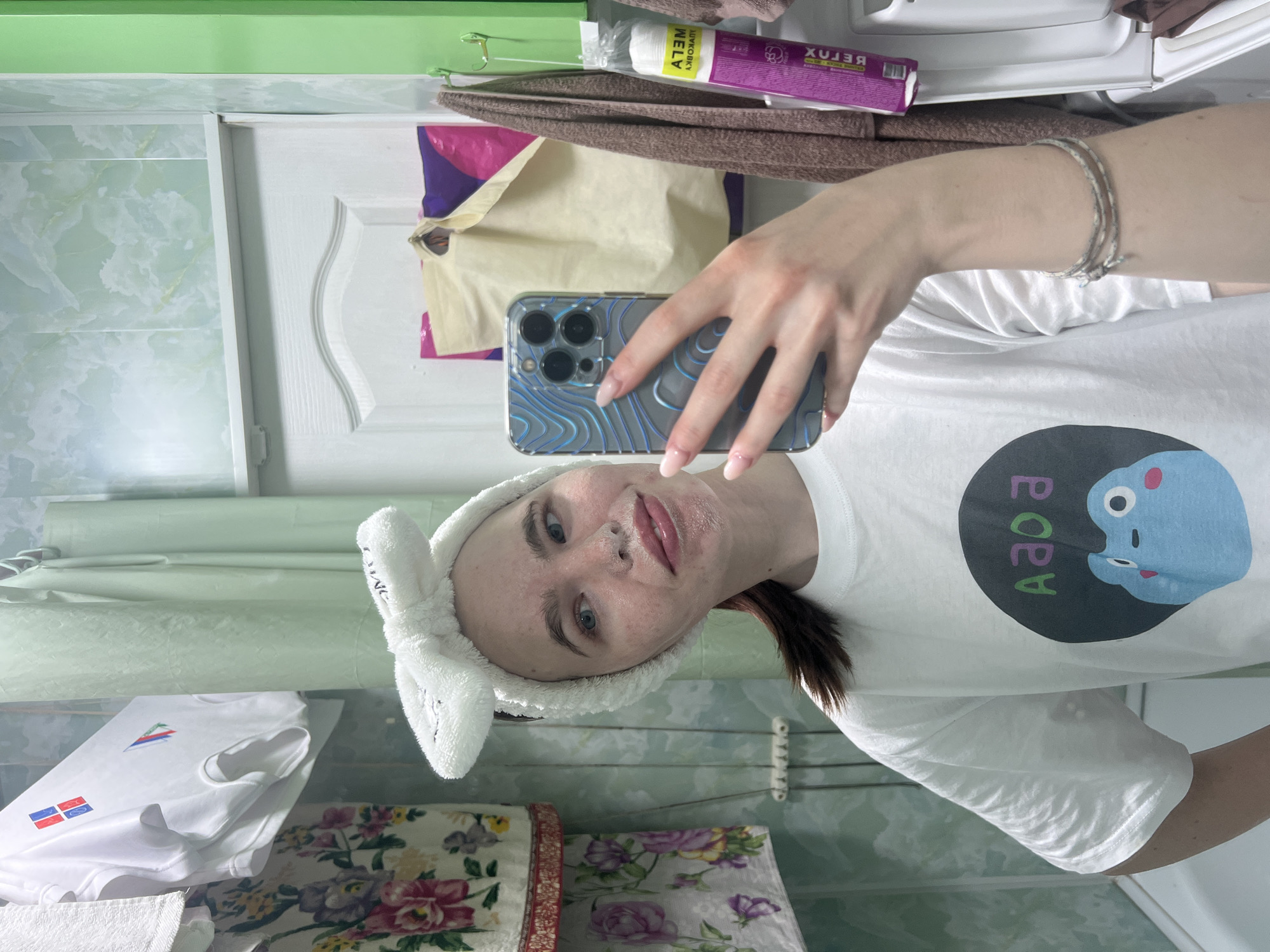 A woman takes a selfie in a bathroom. She wears a white t-shirt with a cartoon character and bunny ear headband. Her phone is held up, obscuring part of her face. The background shows bathroom fixtures, including a towel rack, cabinets, and tiled walls. Other items visible include toiletries, folded towels, and patterned storage boxes. The setting is likely a private residence, potentially in Chaikovsky, Russia.