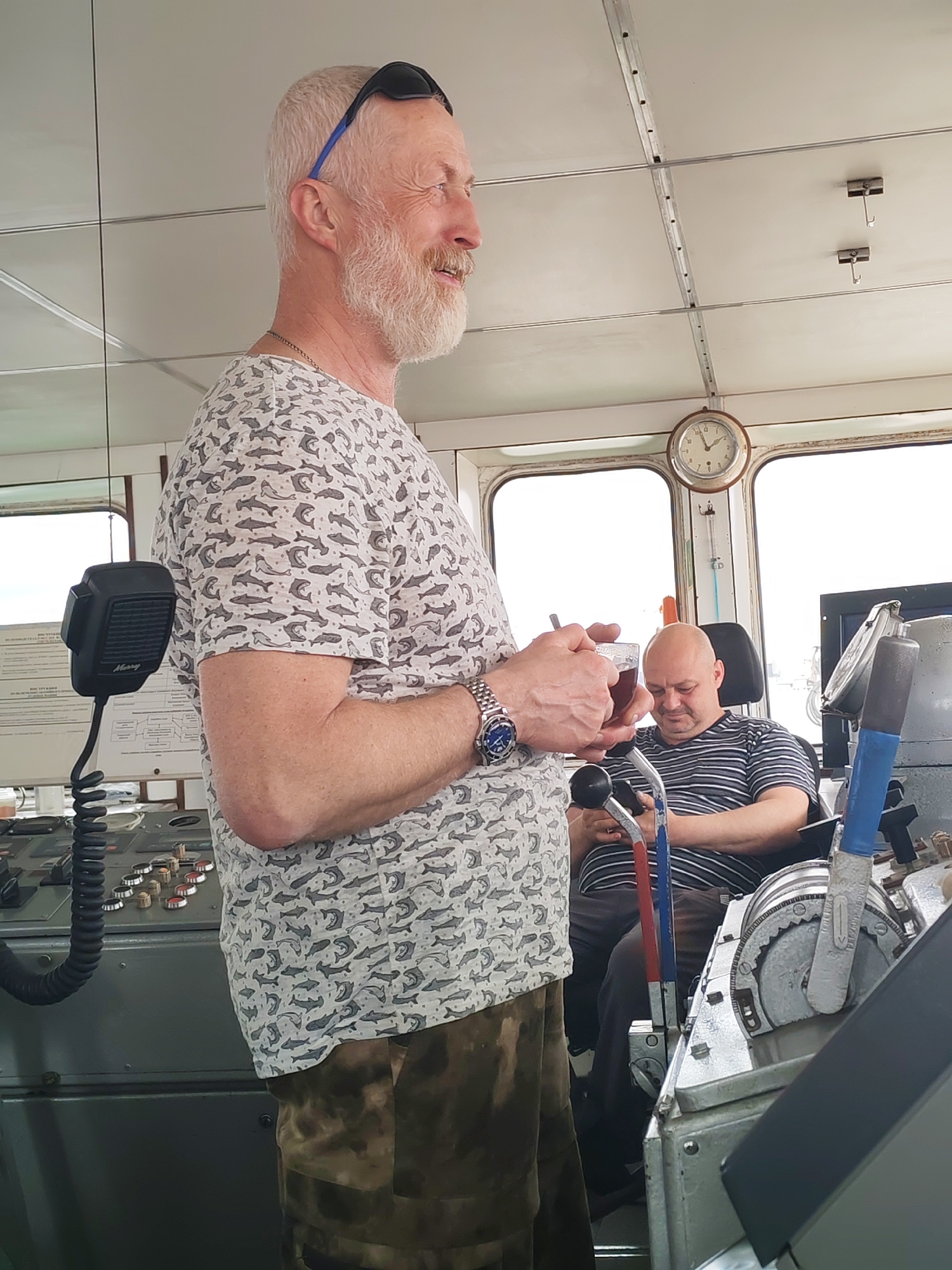 An indoor shot captures two men in what appears to be the bridge or wheelhouse of a ship, set in Городище, Russia.
In the foreground, a fair-skinned man with light gray hair and a prominent beard stands smiling, looking towards the right. He wears sunglasses on his head, a light-colored short-sleeved t-shirt patterned with grey fish, dark patterned trousers, a metal chain, and a wristwatch. He holds a glass cup containing a dark liquid, likely tea, with a spoon or stirrer. To his left, a black "Mercury" brand microphone with a coiled cord is mounted on a control console featuring various buttons and dials.
In the mid-ground, a bald man with fair skin is seated in a black chair, wearing a striped short-sleeved t-shirt. He looks down, engrossed in a small device, possibly a mobile phone. Behind him, parts of the ship's controls, including large red, blue, and gray levers, are visible.
The setting features large windows in the background, revealing an overcast sky, suggesting daytime. A round wall clock mounted above the windows shows the time to be approximately 11:30. The ceiling is white and paneled with metal strips and hooks. On the left, papers with visible but illegible Cyrillic text and a diagram are attached to a board.
The scene depicts a casual moment, possibly a break, within the utilitarian environment of a vessel's command deck. The standing man appears relaxed and content, while the seated man is focused on his device.