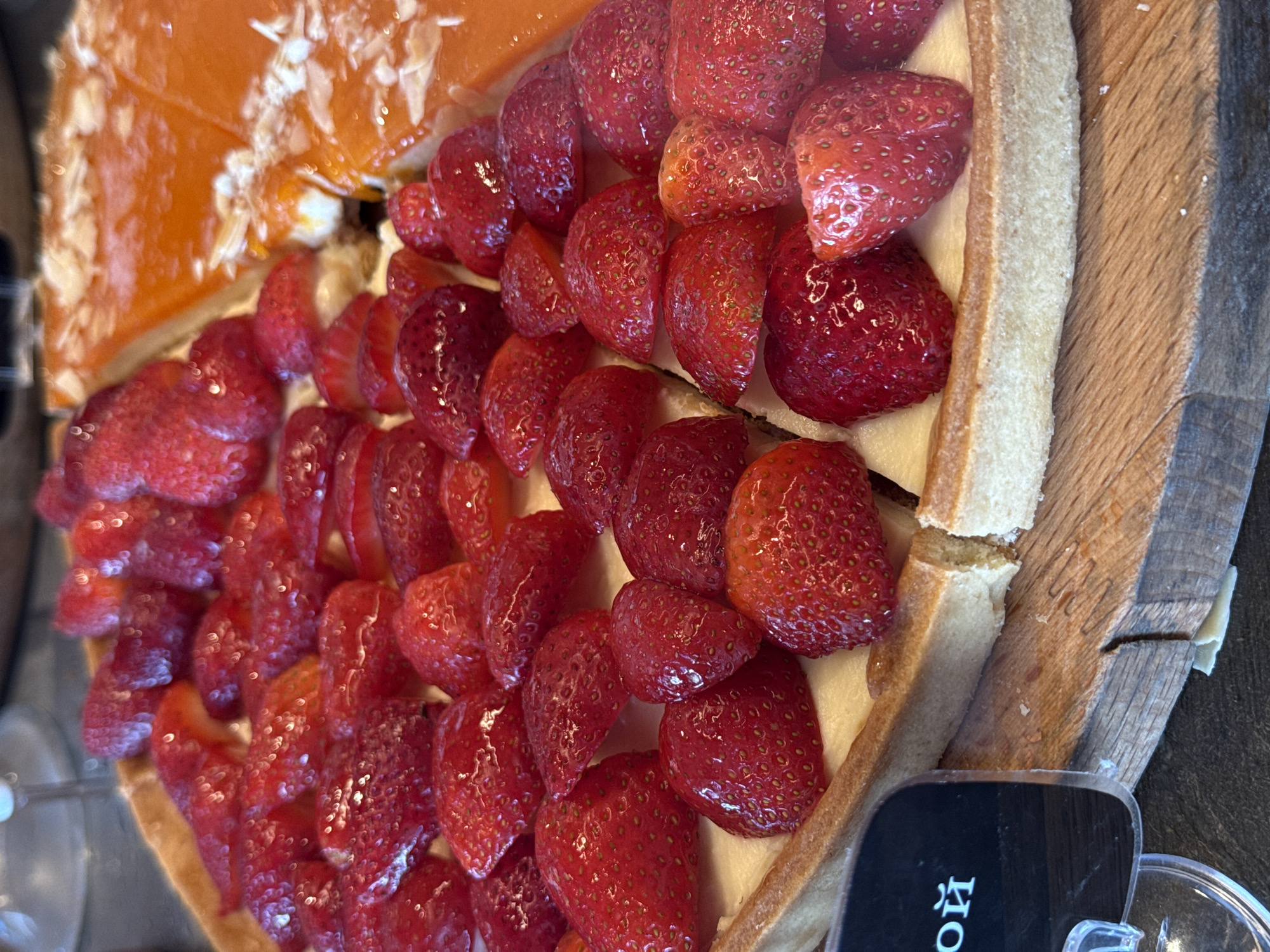 The image displays two enticing dessert items, likely tarts or cakes, presented side-by-side on a rustic wooden surface.
The most prominent dessert, occupying the right and lower portions of the frame, is a large strawberry tart. It features numerous vibrant, fresh red strawberries, mostly halved, arranged meticulously on a creamy white or pale yellow base, which appears to be a custard or cream cheese filling. The strawberries have a glossy sheen, suggesting they might be coated with a light glaze. The tart's golden-brown crust is visible along its right edge, indicating a firm, baked texture.
Partially visible in the upper left of the image is a slice of another dessert. This item has a smooth, glossy orange-colored top layer, possibly an apricot or citrus glaze, adorned with scattered white flakes, which could be almond slivers or coconut.
The desserts are arranged on a wooden board or platter, showcasing a distinct grain pattern that contributes to an artisanal presentation. A small, black rectangular label or sign is partially visible in the bottom right corner of the image, placed next to the desserts.
The activity depicted is the display of these visually appealing desserts, characteristic of a cafe, bakery, or dessert shop, likely prepared for sale or consumption.
On the black label, the Cyrillic letters "ЙО" are clearly visible in white, bold font. This is likely part of a larger word, such as a product name or ingredient description, fitting the context of Sochi, Russia. No people are visible in the image, and the time of day and weather are not discernible.